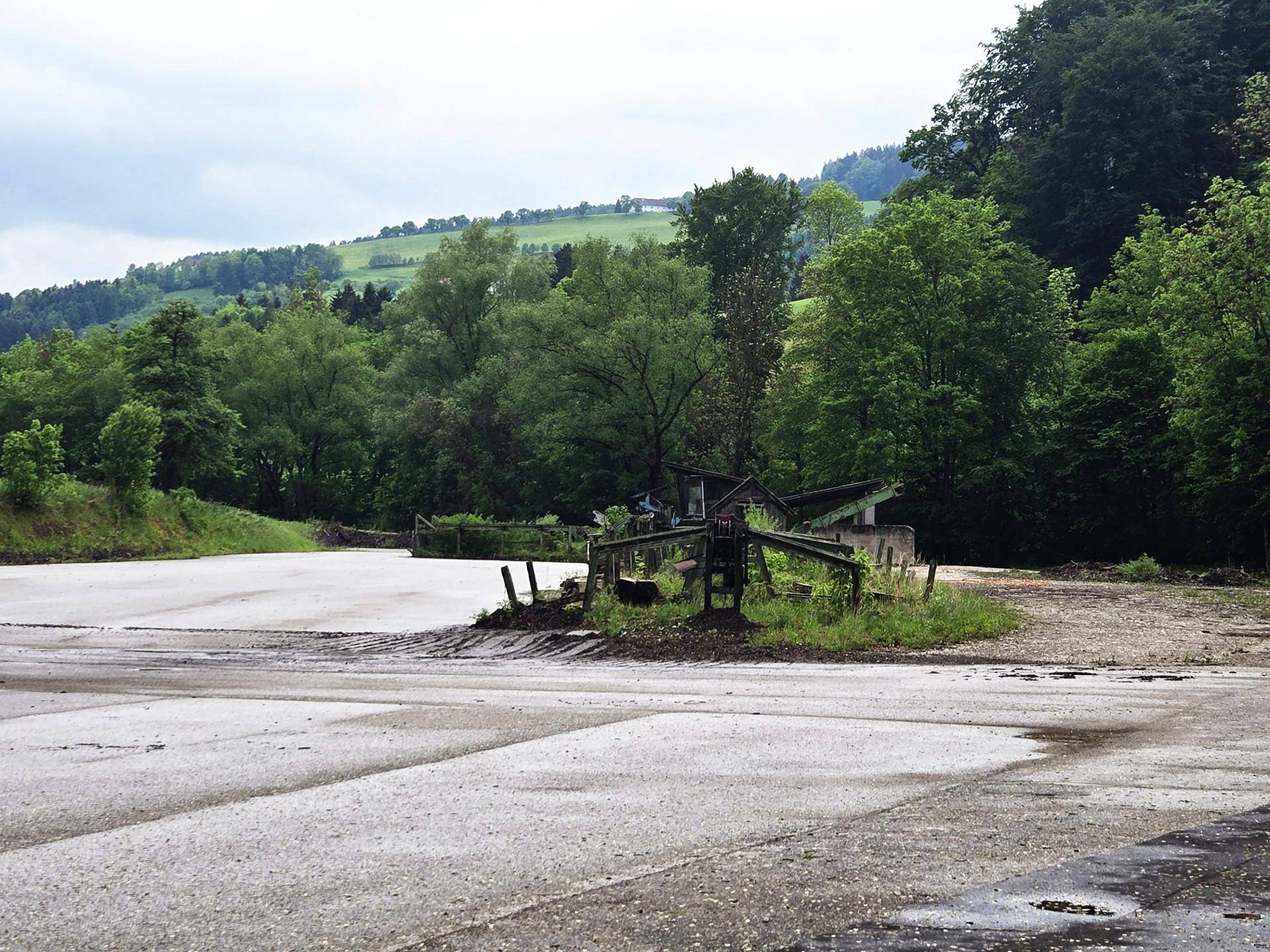 Weitläufige Außenfläche mit Schotter und Asphalt, umgeben von üppiger grüner Landschaft und bewaldeten Hügeln.