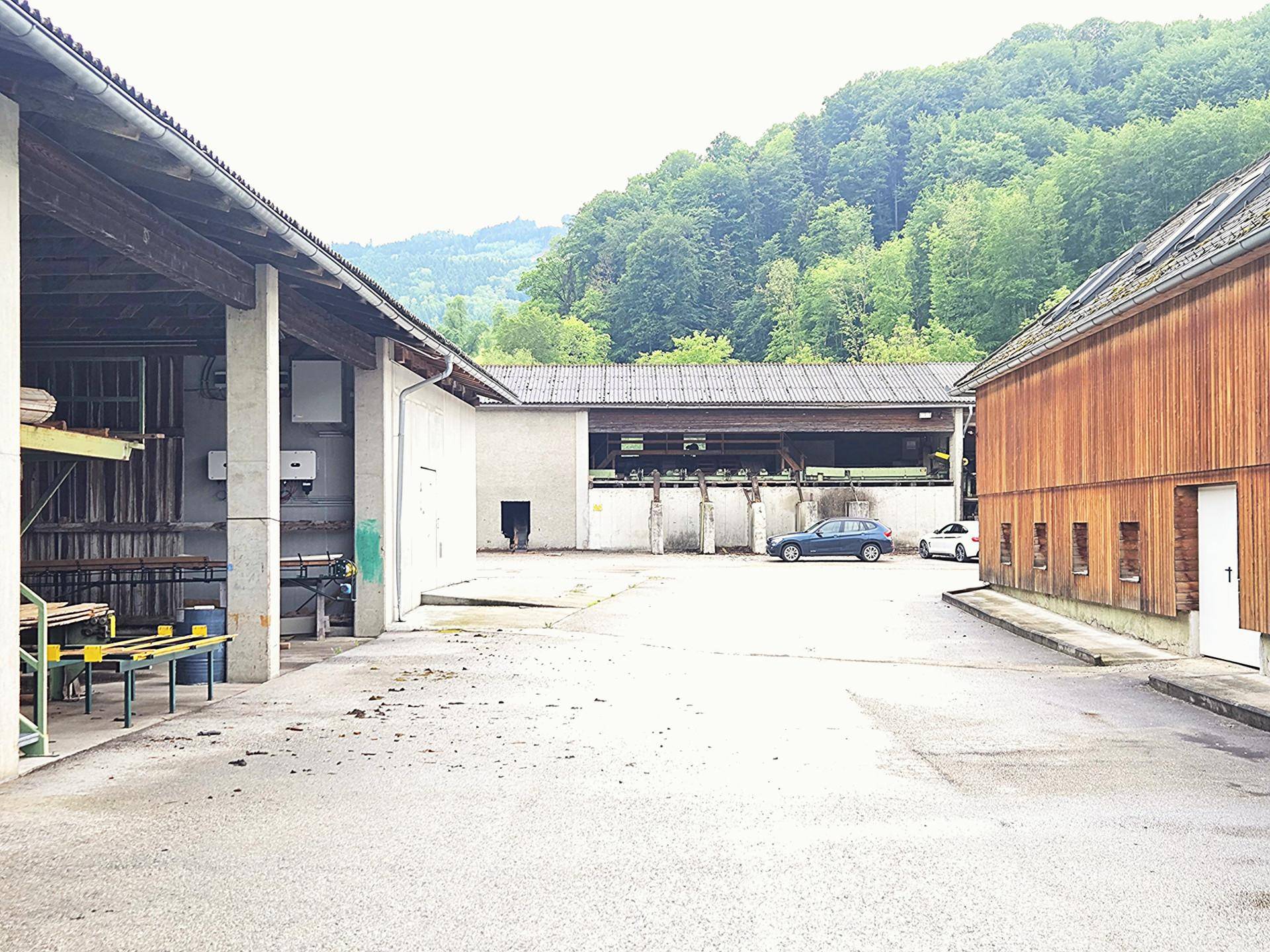 Innenhof zwischen mehreren Gebäuden mit Holz- und Betonfassaden, im Hintergrund parken Autos und grüne Hügel.