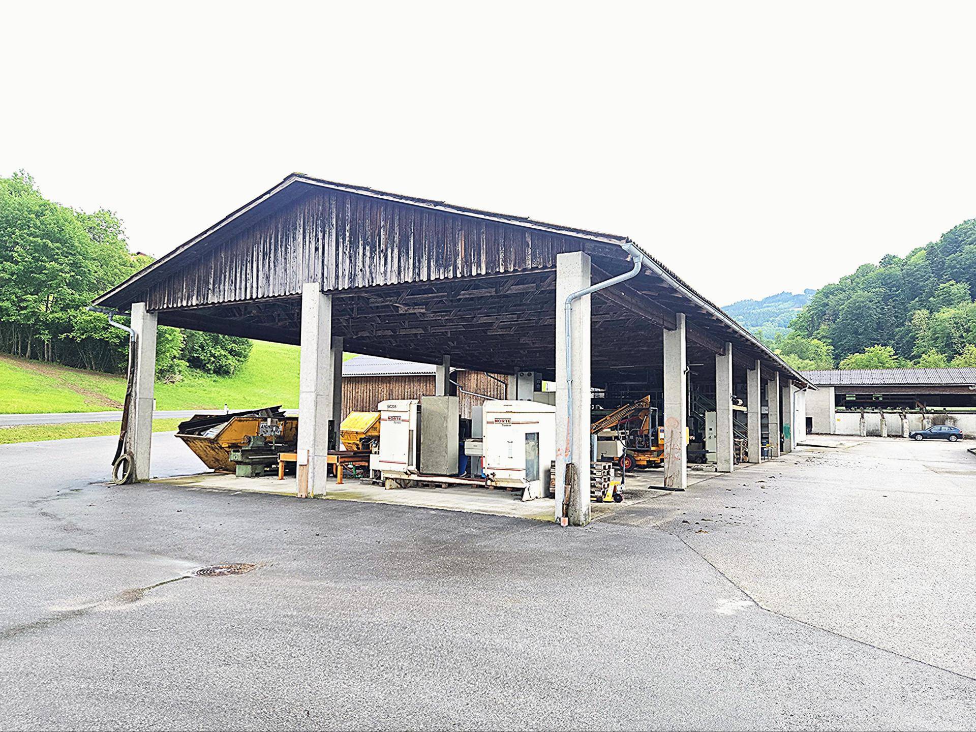 Große offene Halle mit Holzdach und Betonpfeilern, darunter verschiedene Maschinen und Geräte gelagert.