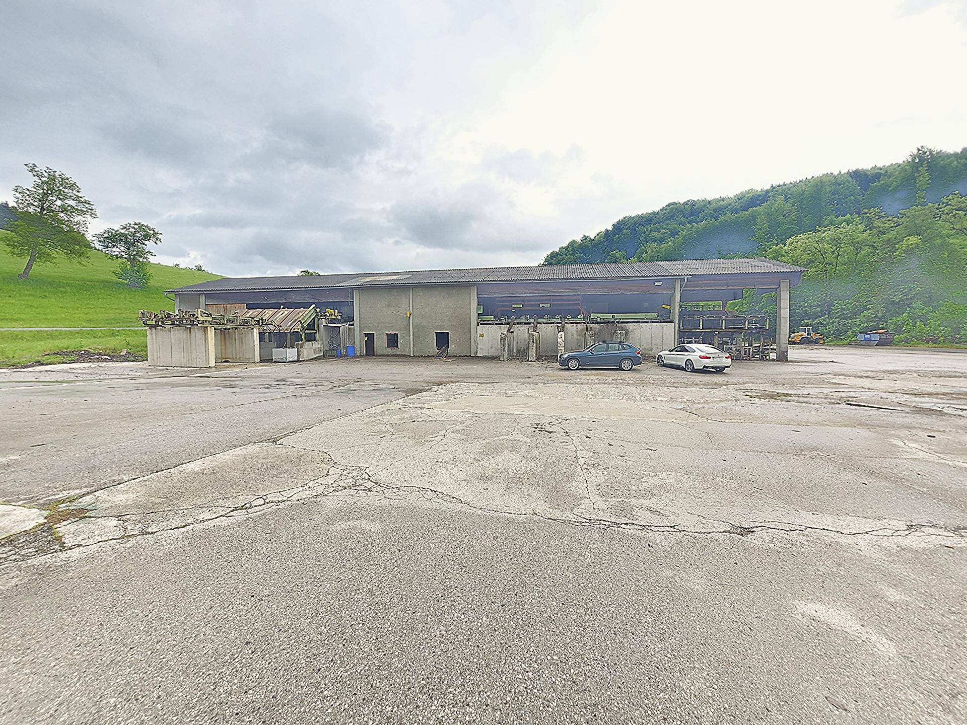 Langes, offenes Gebäude mit Flachdach und Betonstruktur auf einer großen asphaltierten Fläche, davor parken Autos.