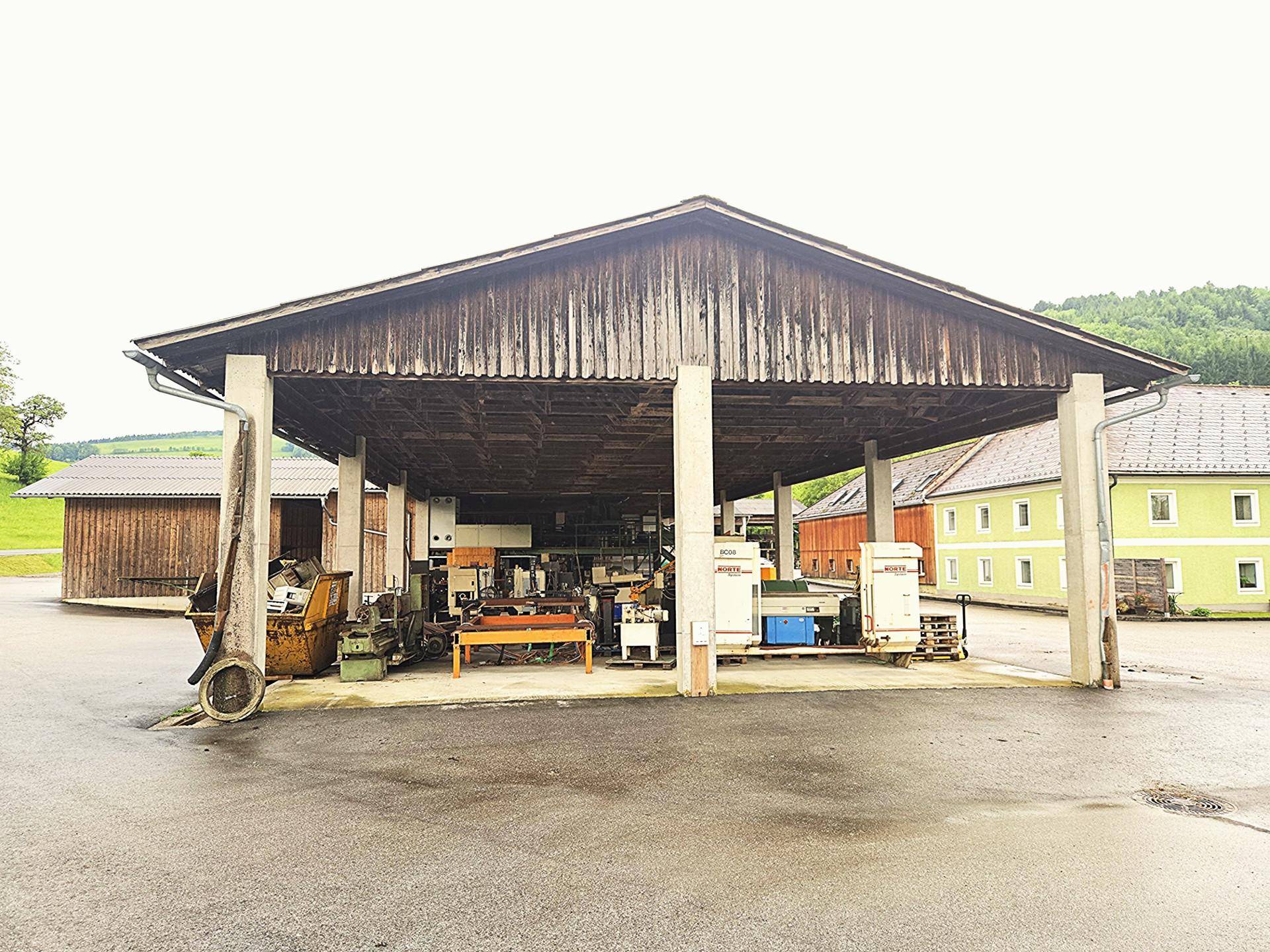 Offene Halle mit Holzdach und Betonpfeilern, in der Maschinen gelagert sind, im Hintergrund ein traditionelles Haus.