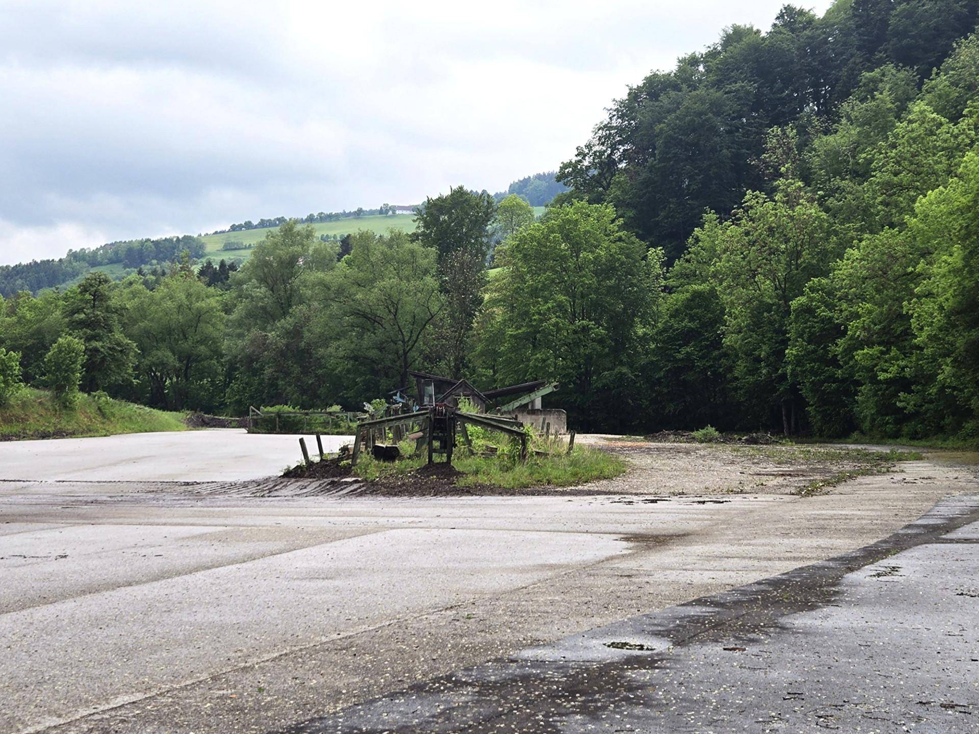 Weitläufige Außenfläche mit Schotter und Asphalt, umgeben von üppiger grüner Landschaft und bewaldeten Hügeln.