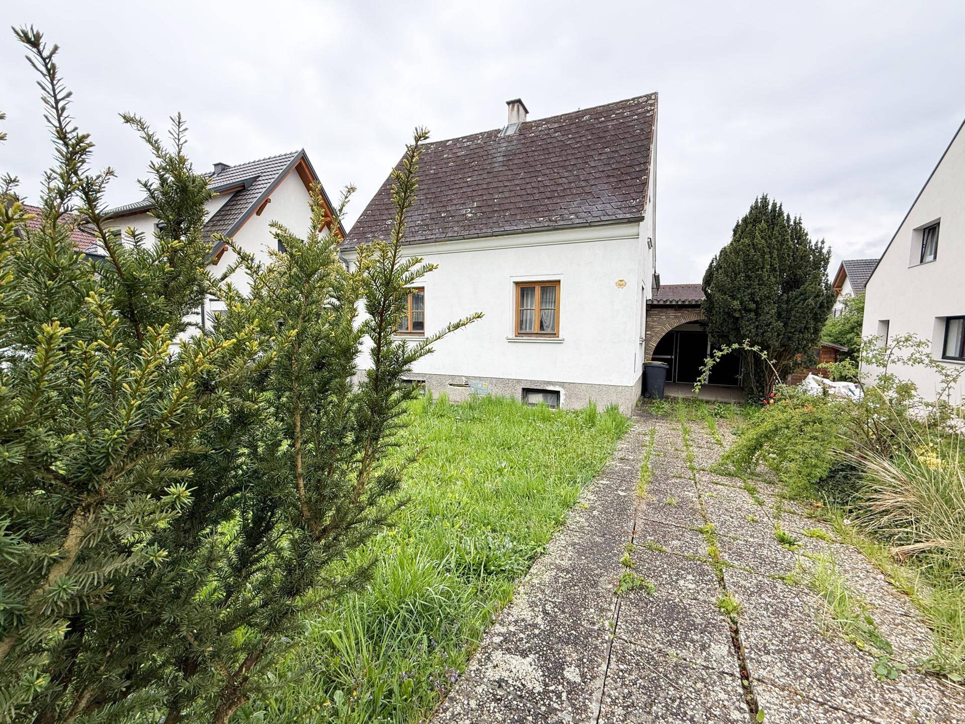 Seitenansicht eines weißen Hauses mit Satteldach und einem zugewachsenen Weg im Gartenbereich.