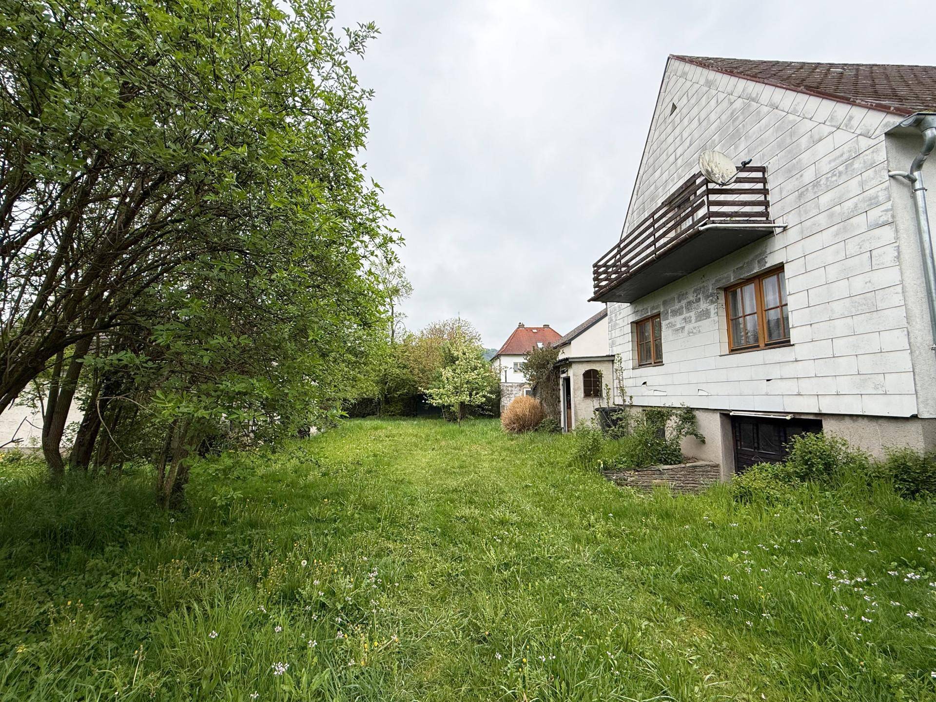 Rückansicht eines Hauses mit Balkon und einem großen, verwilderten Gartenbereich unter bewölktem Himmel.