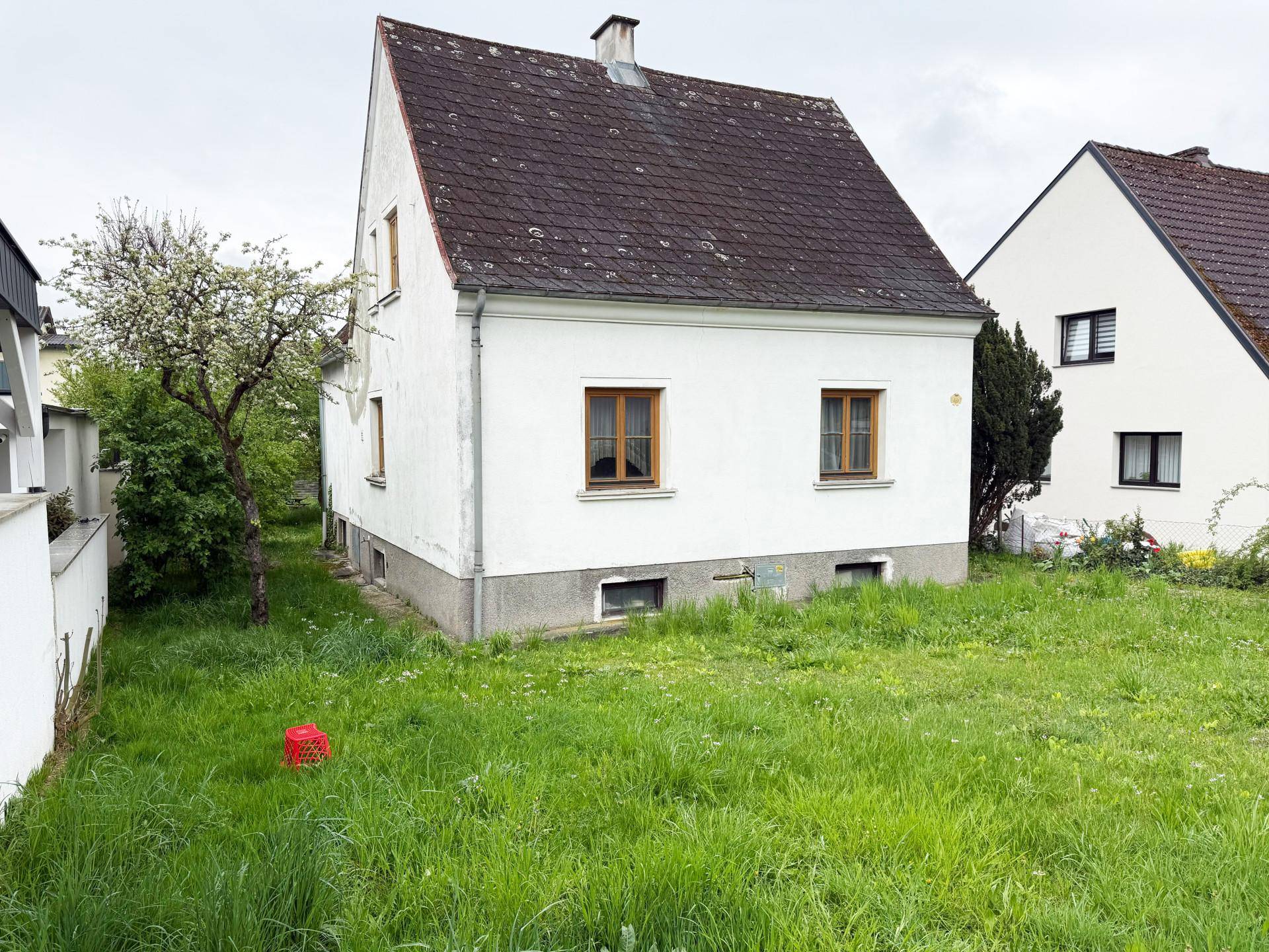 Freistehendes Einfamilienhaus mit weißer Fassade und großem Gartenbereich, der Pflege benötigt.
