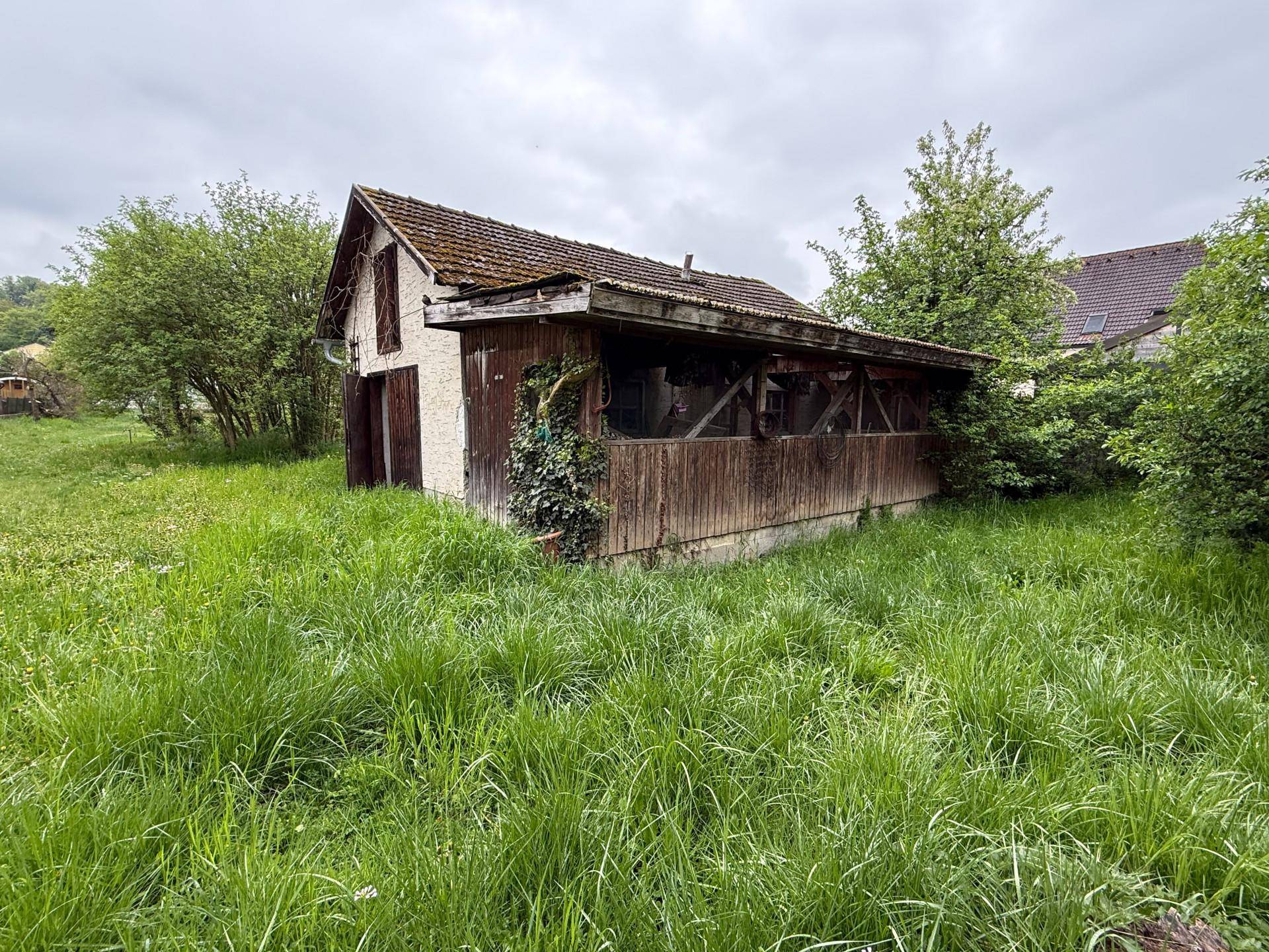 Altes Nebengebäude mit Holzanbau und verwildertem Garten, das Renovierungspotenzial bietet.