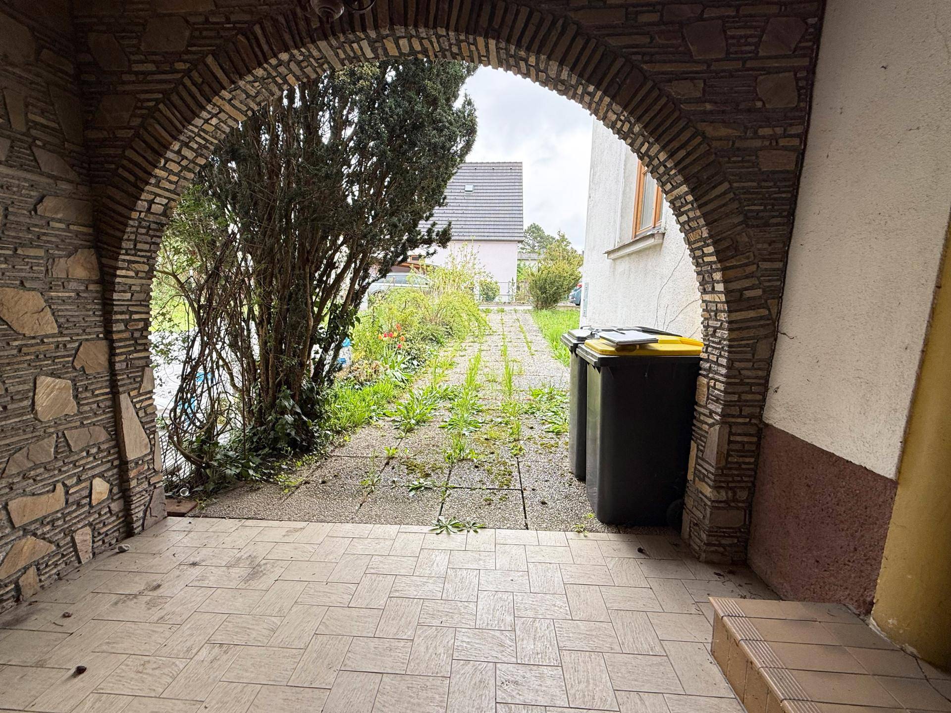 Überdachter Eingangsbereich mit einem Steinbogen und Blick auf einen verwachsenen Weg im Freien.