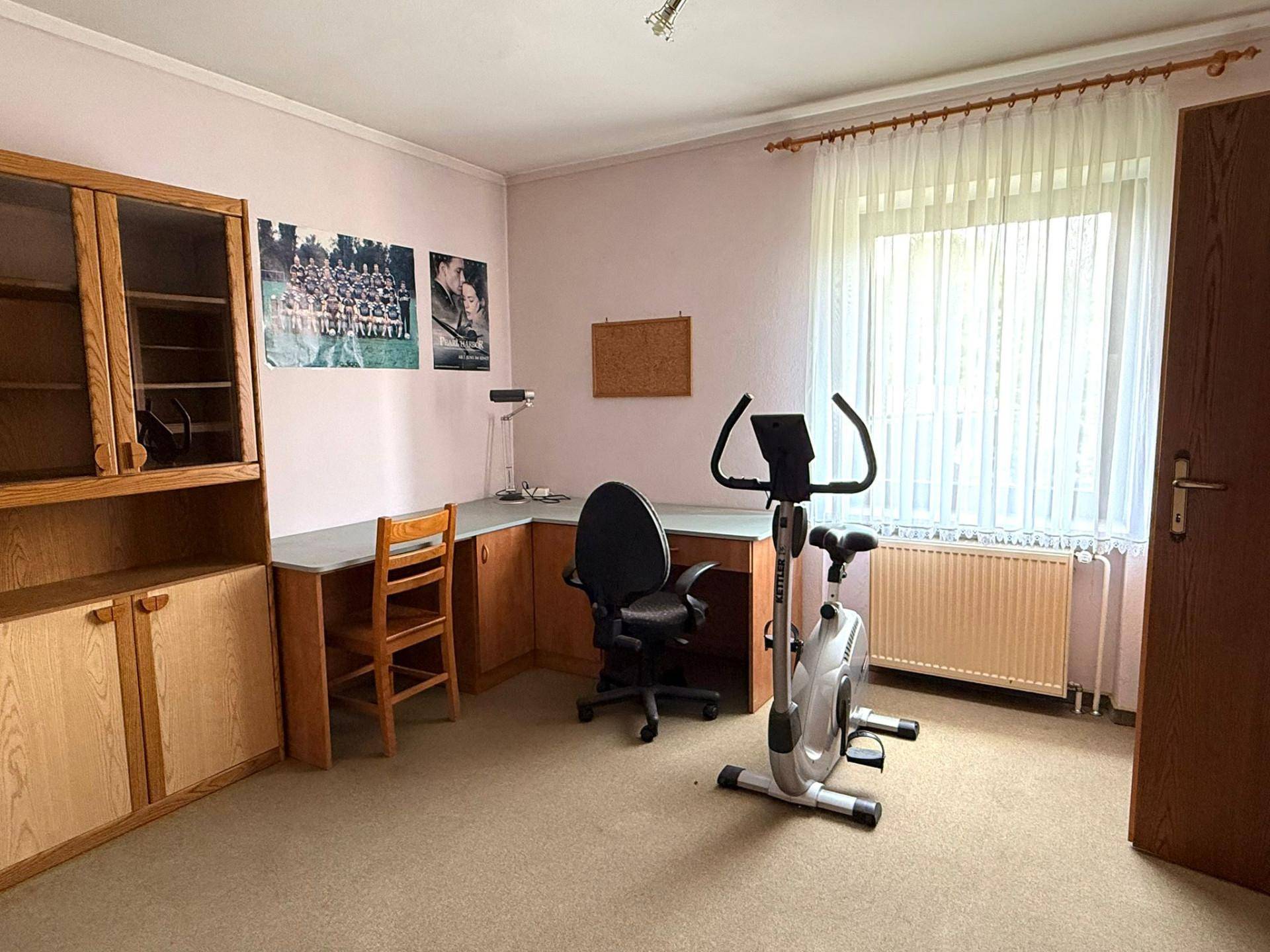 Geräumiges Zimmer mit Schreibtisch, Bücherregal und Heimtrainer am Fenster.