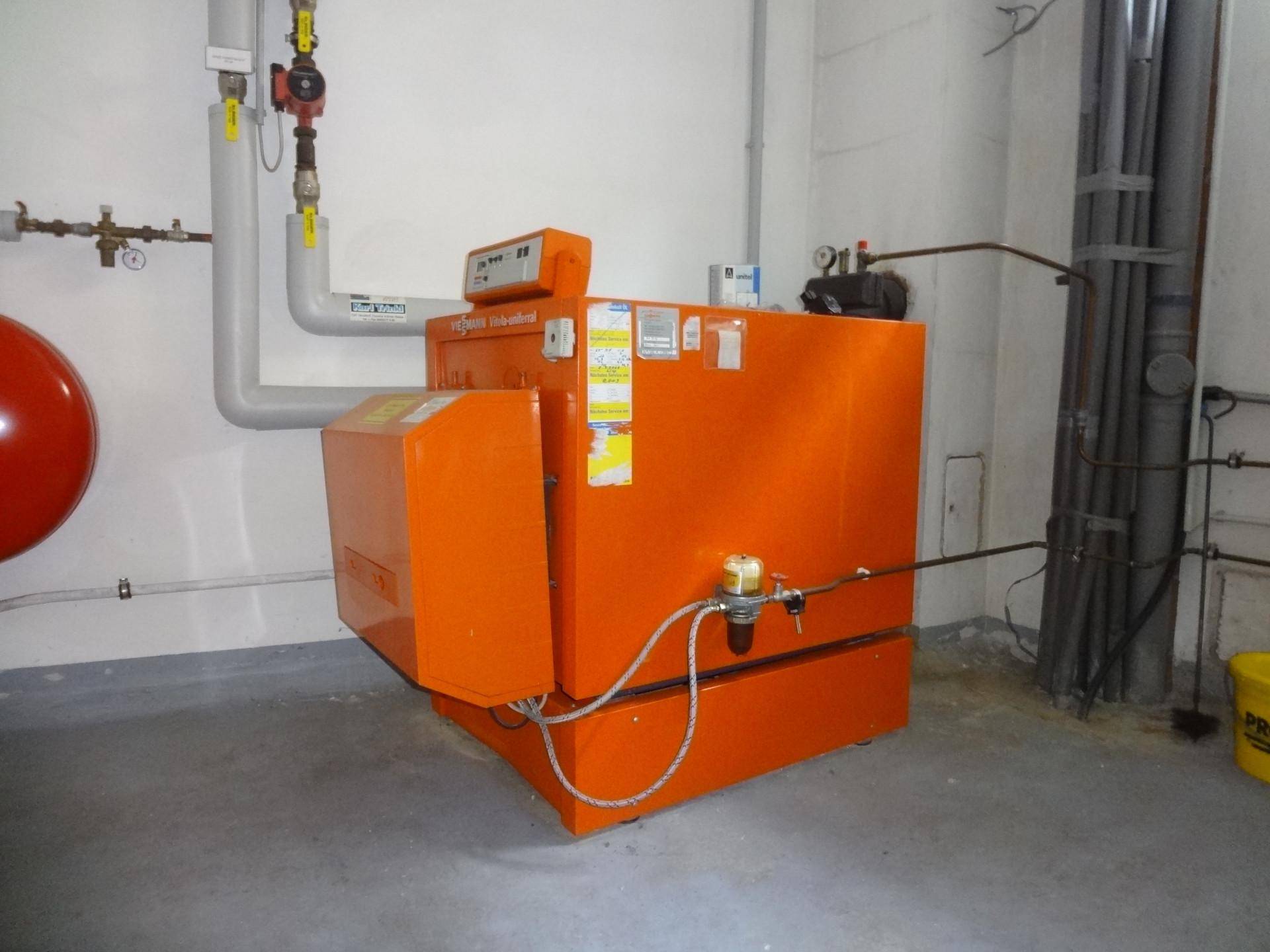 Ein Heizraum mit einem großen orangefarbenen Heizkessel und sichtbaren Rohrleitungen an der Wand.
