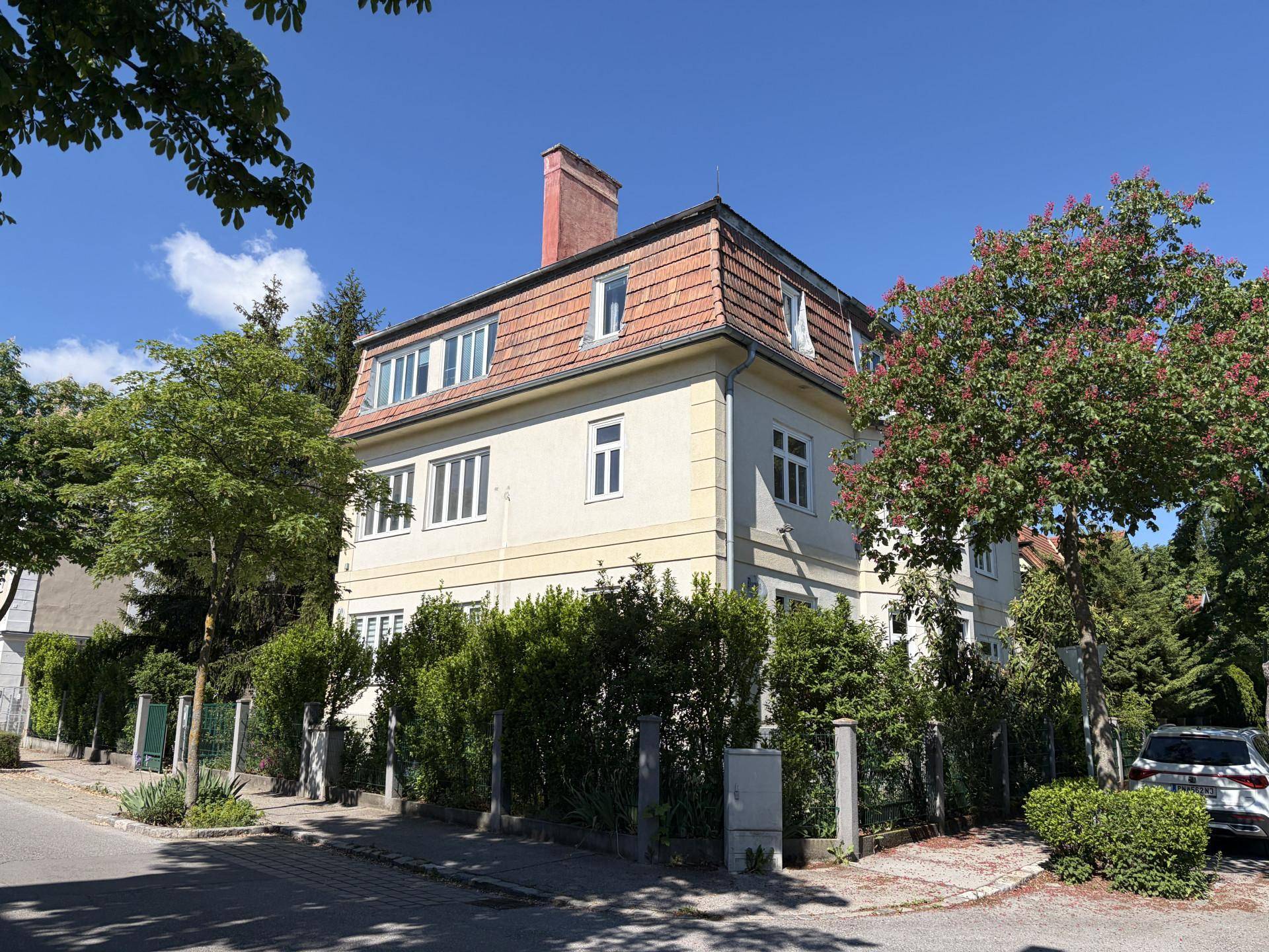Historisches Gebäude mit Mansarddach und gepflegter Fassade, umgeben von grünen Hecken.