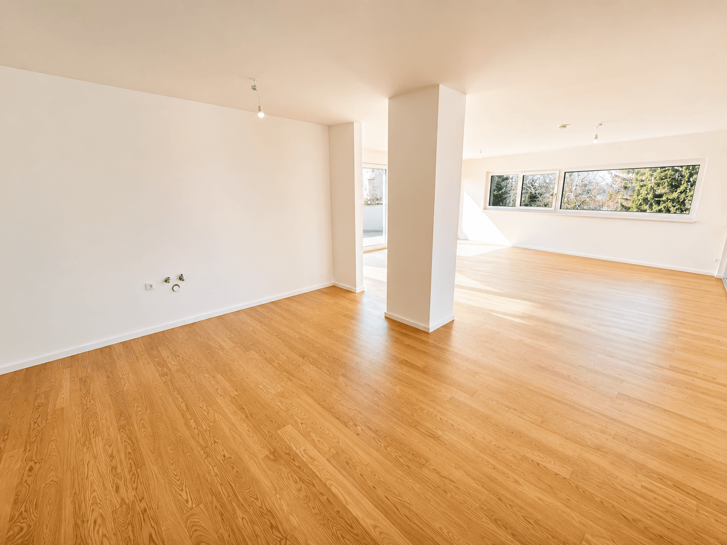 Heller, offener Wohnbereich mit Holzboden, großen Fenstern und Zugang zu einem Balkon im Hintergrund.