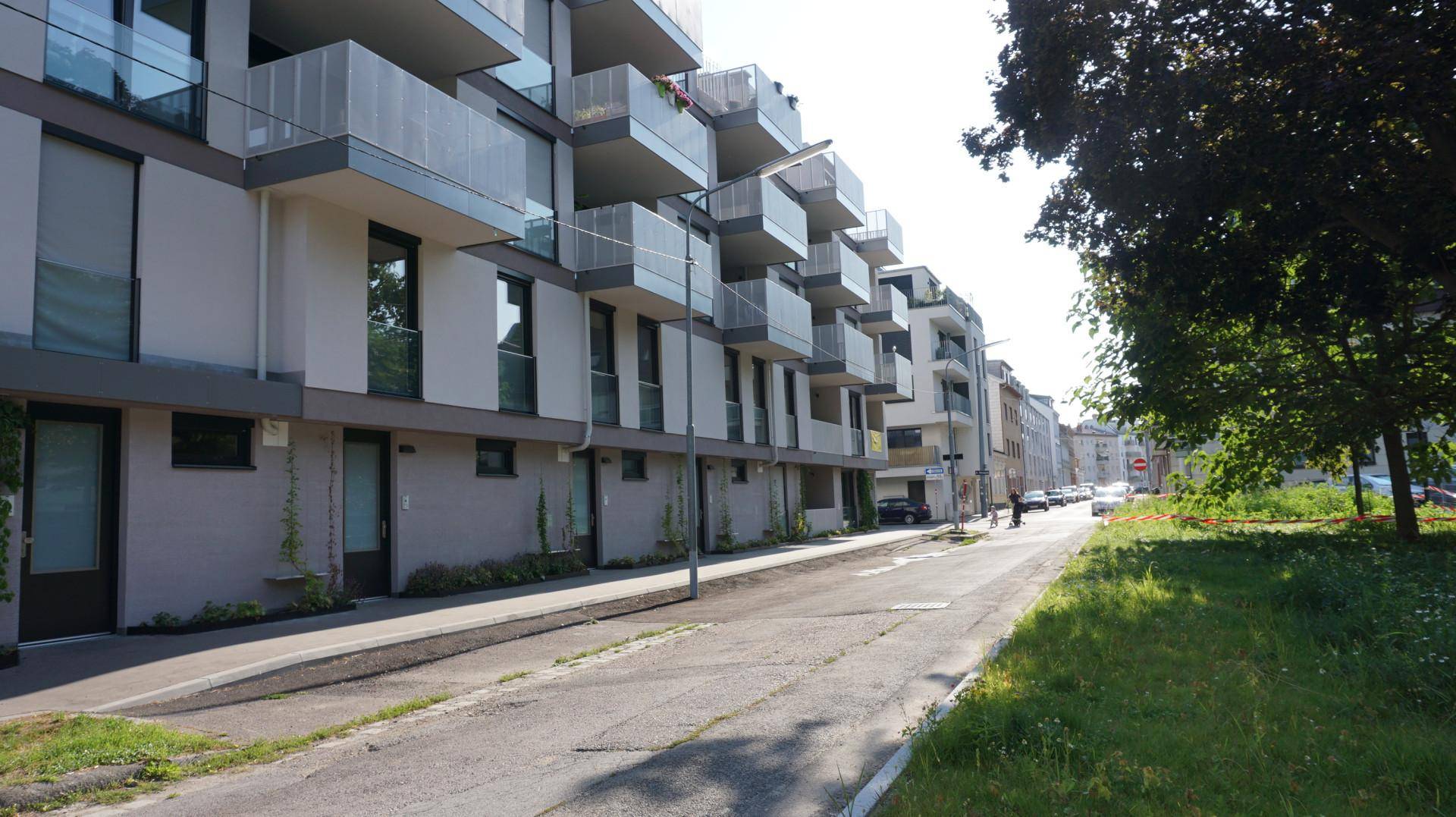 Seitenansicht eines modernen Wohngebäudes entlang einer ruhigen Straße mit Bäumen und Grünflächen.