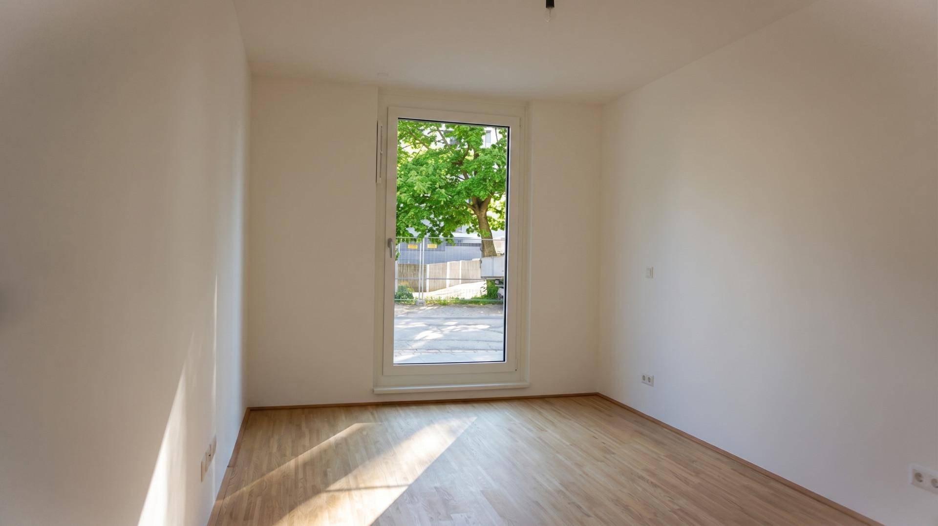 Leeres Zimmer mit Holzboden und einem großen Fenster, das viel Tageslicht hereinlässt.