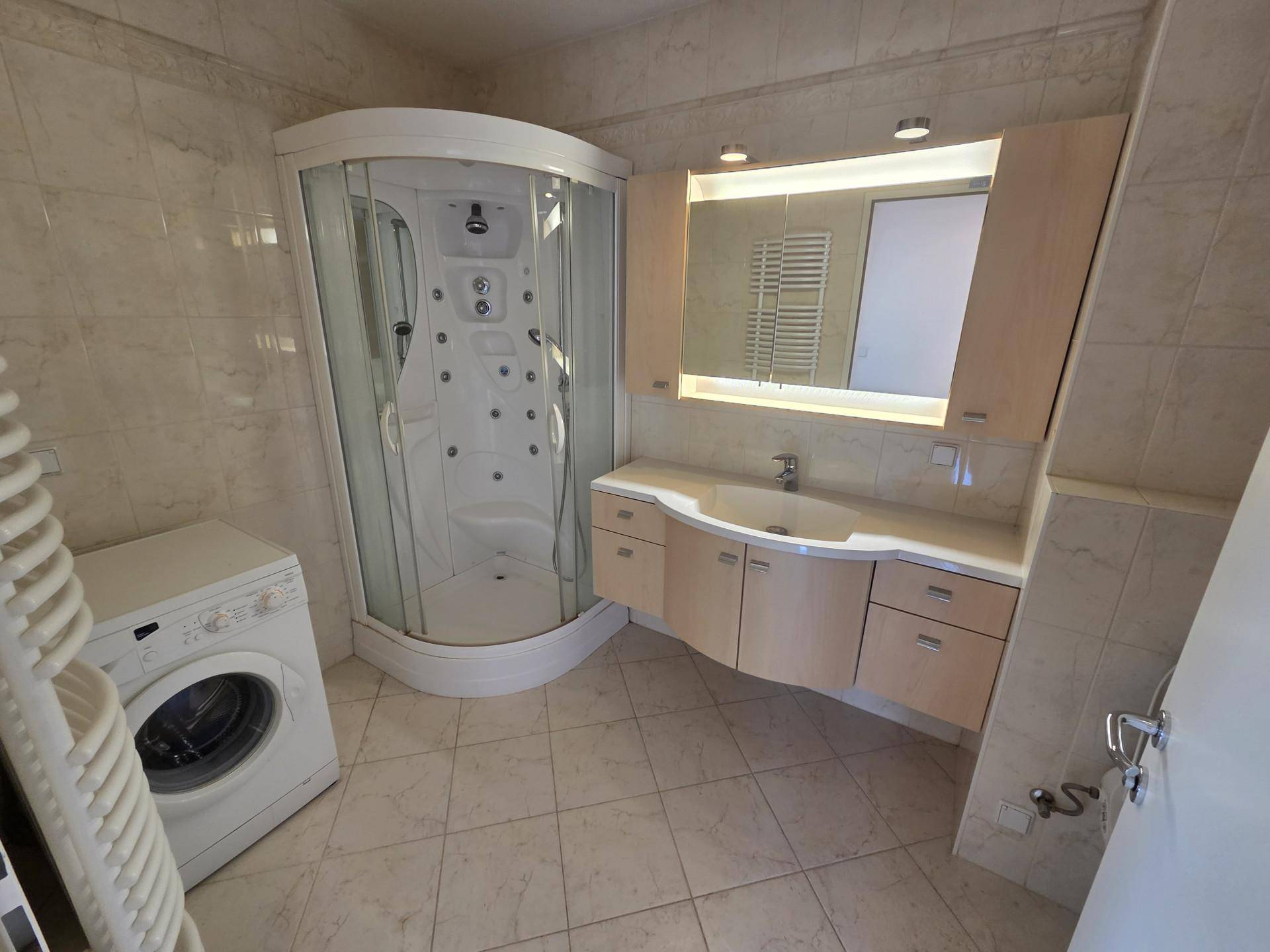 Badezimmer mit Eckduschkabine, Waschtisch und Waschmaschine, hell gefliest.