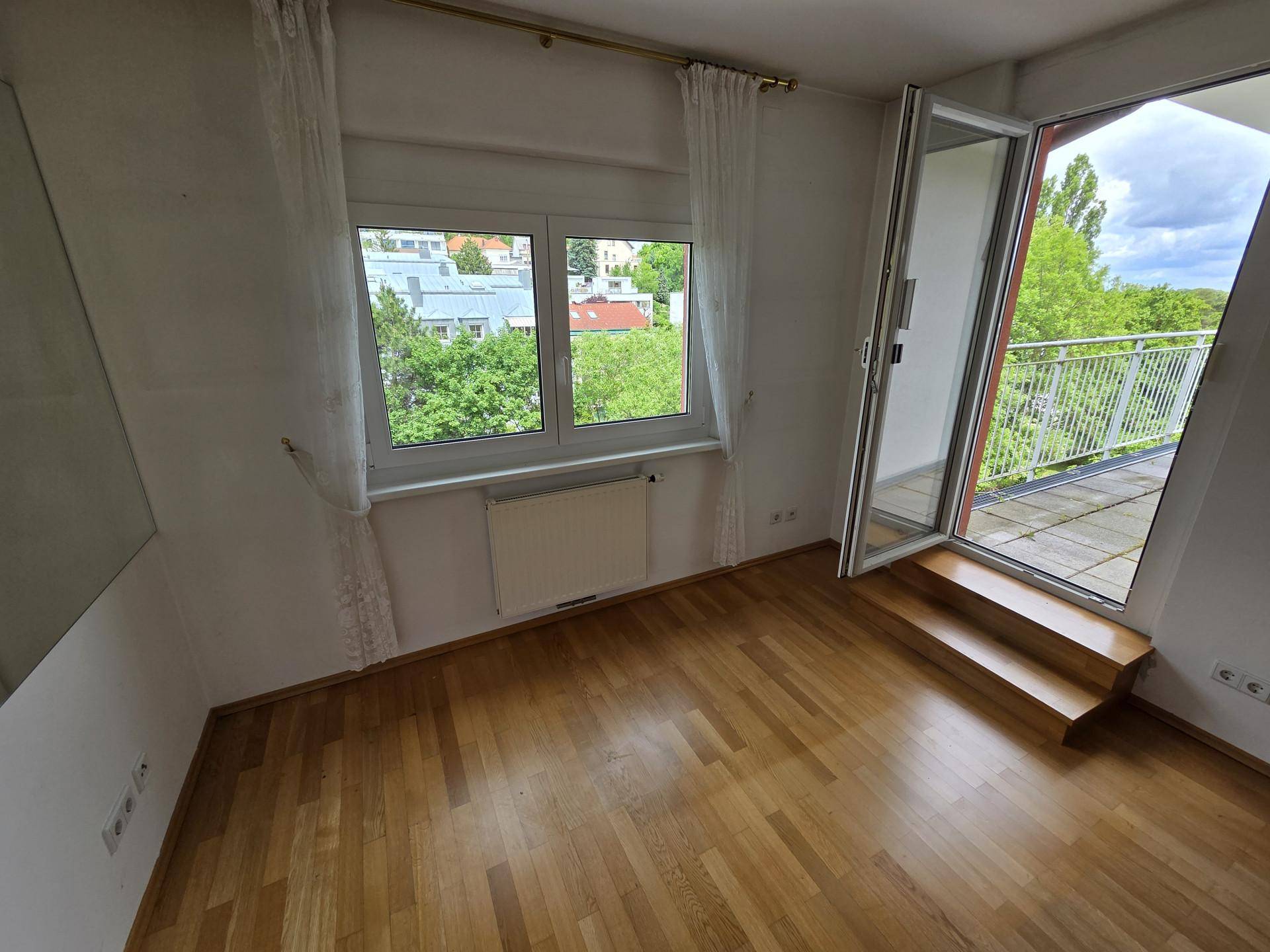 Helles Zimmer mit Parkettboden, Fenster und Zugang zu einem Balkon oder einer Terrasse.