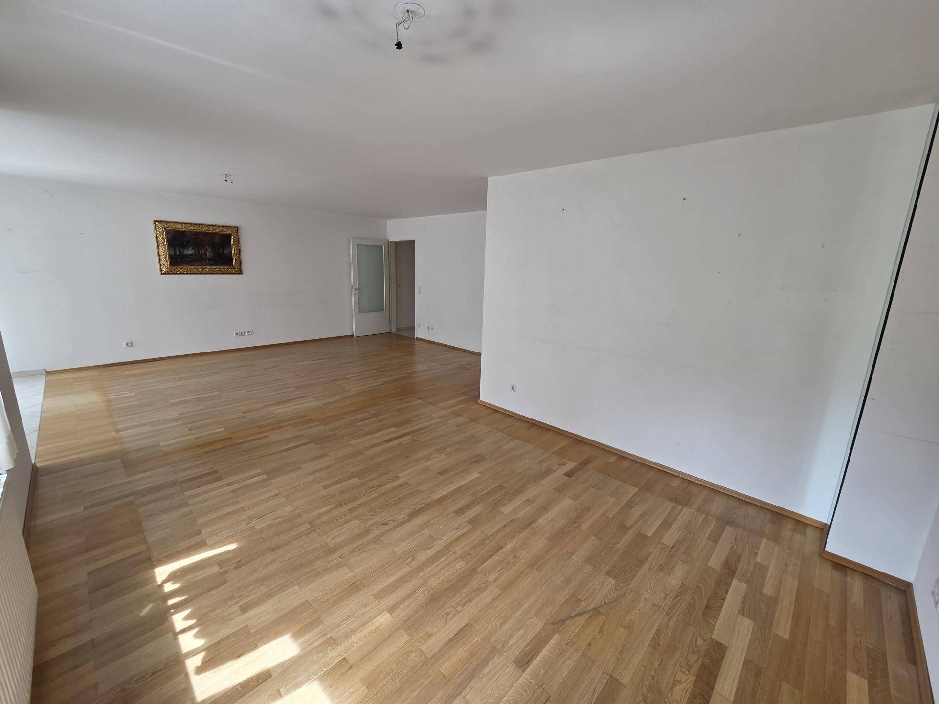 Geräumiges leeres Zimmer mit Parkettboden, weißen Wänden und einem dekorativen Bild.