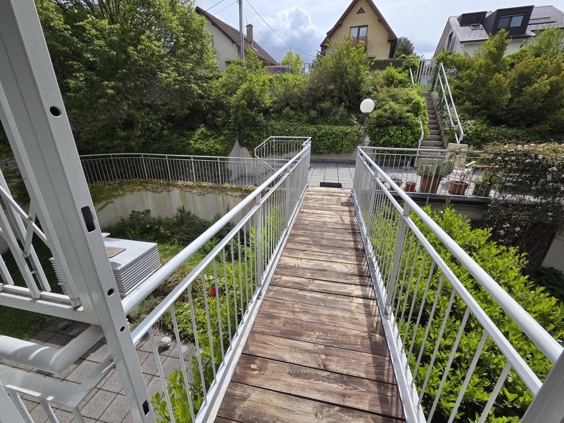 Hölzerner Steg mit weißen Geländern, der zu einer gefliesten Terrasse und Grünflächen führt.