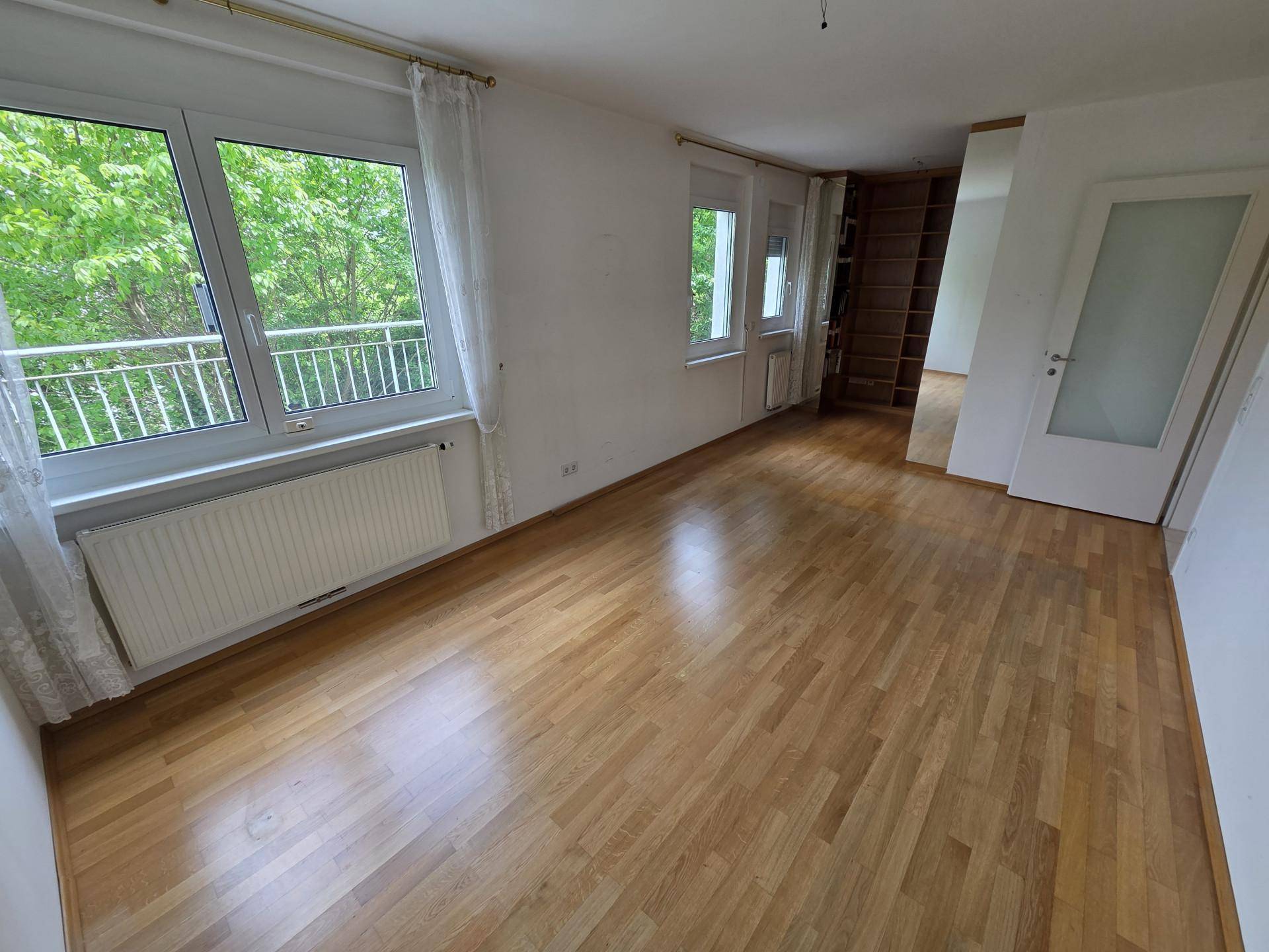 Helles Zimmer mit Parkettboden, Fenstern und einem integrierten Bücherregal.