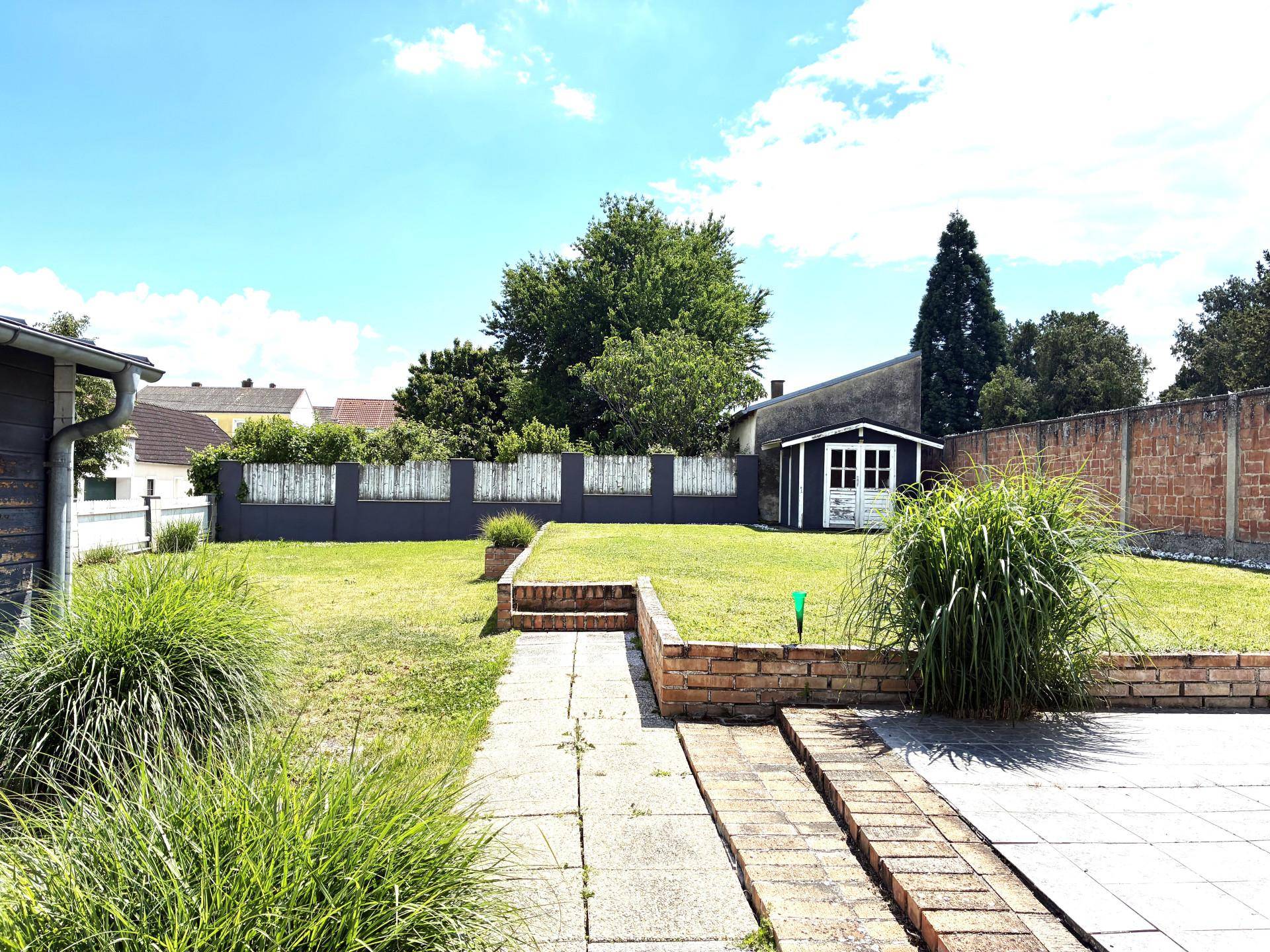 Gepflegter Garten mit Rasenfläche, Gartenhaus und gepflastertem Weg unter blauem Himmel.