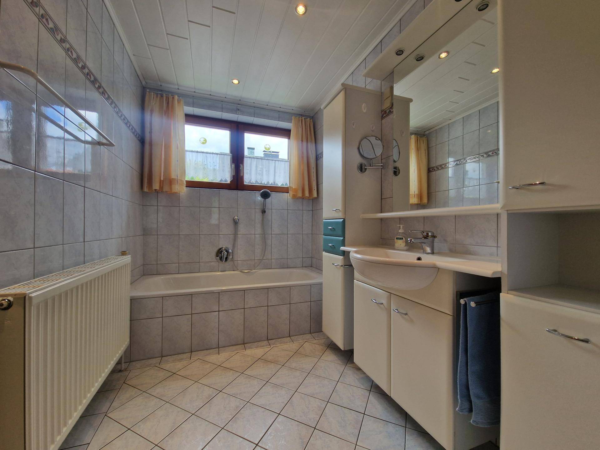 Gefliestes Badezimmer mit Badewanne, Duschfunktion, Waschtisch und Fenster.