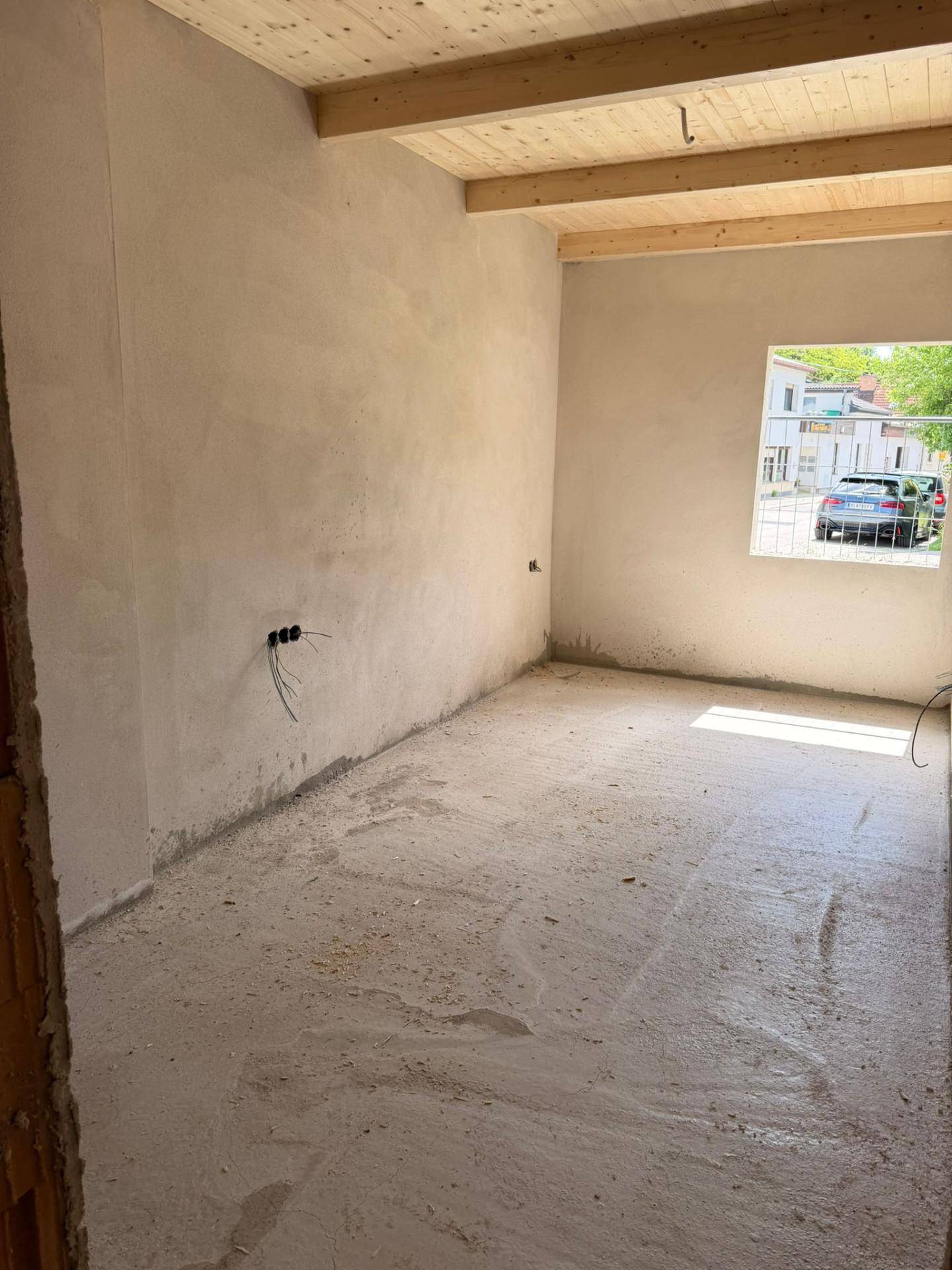 Unfertiger Raum mit Holzbalkendecke, Betonboden und einem Fenster zur Straße.
