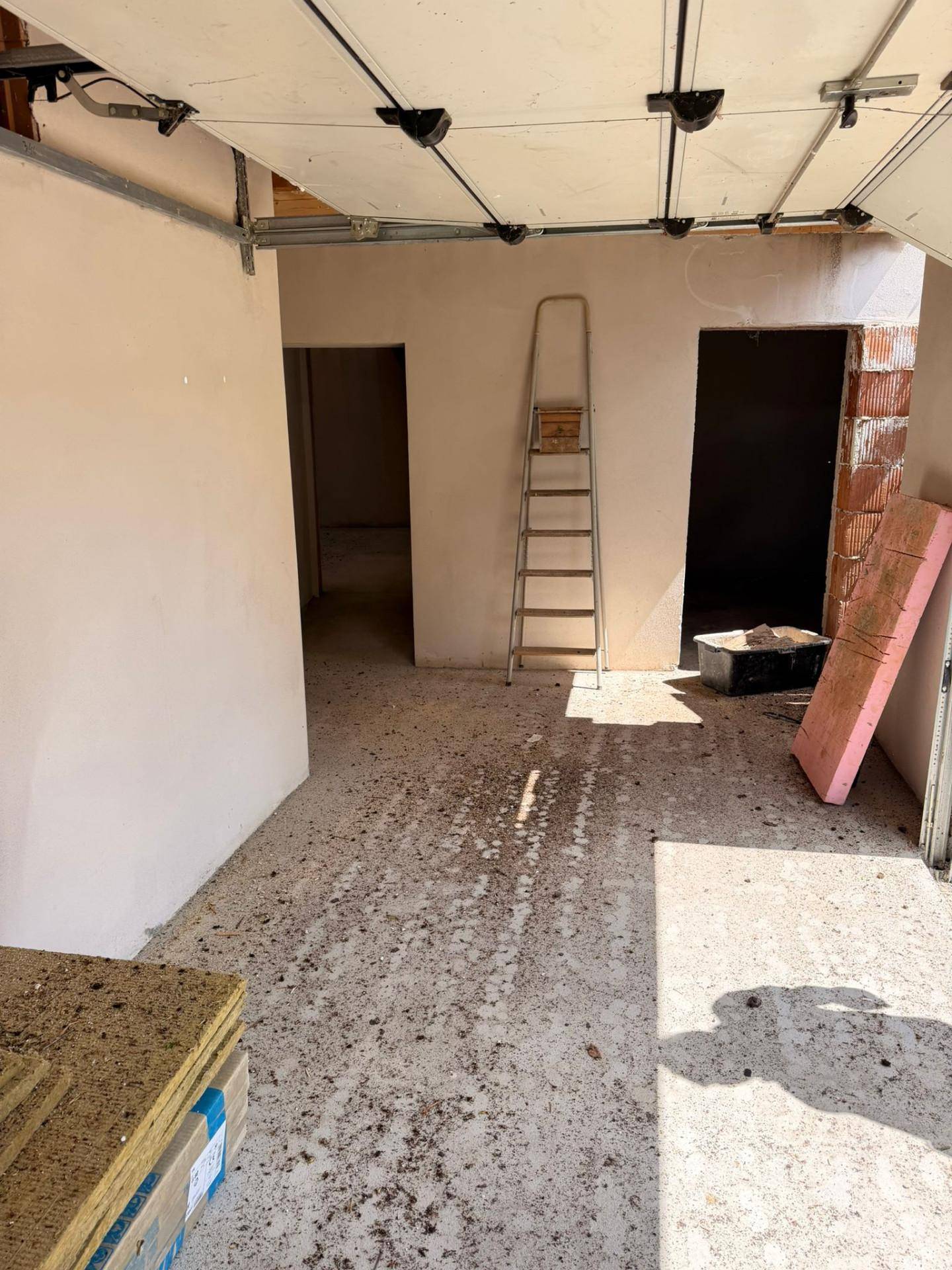 Unfertige Garage mit Betonboden, Rolltor und Zugang zu weiteren Räumen.