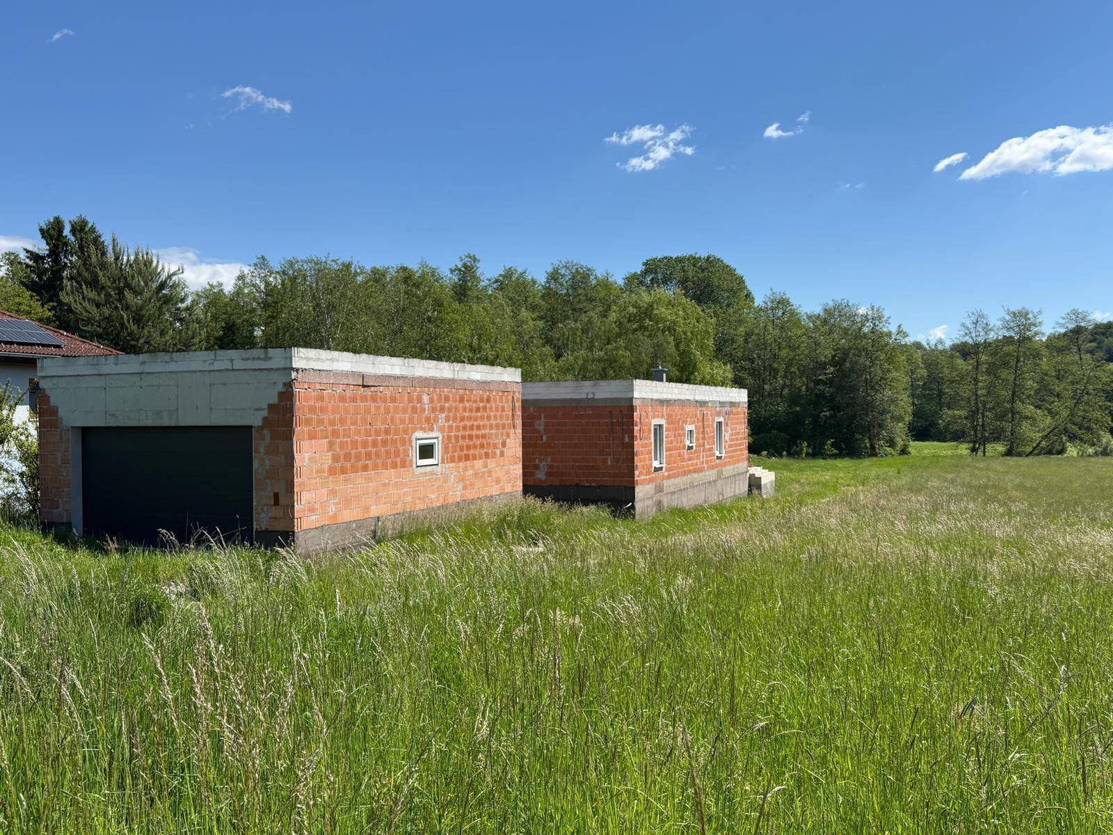Zwei im Bau befindliche Gebäude, eines davon mit Garagentor, inmitten einer weitläufigen grünen Landschaft.