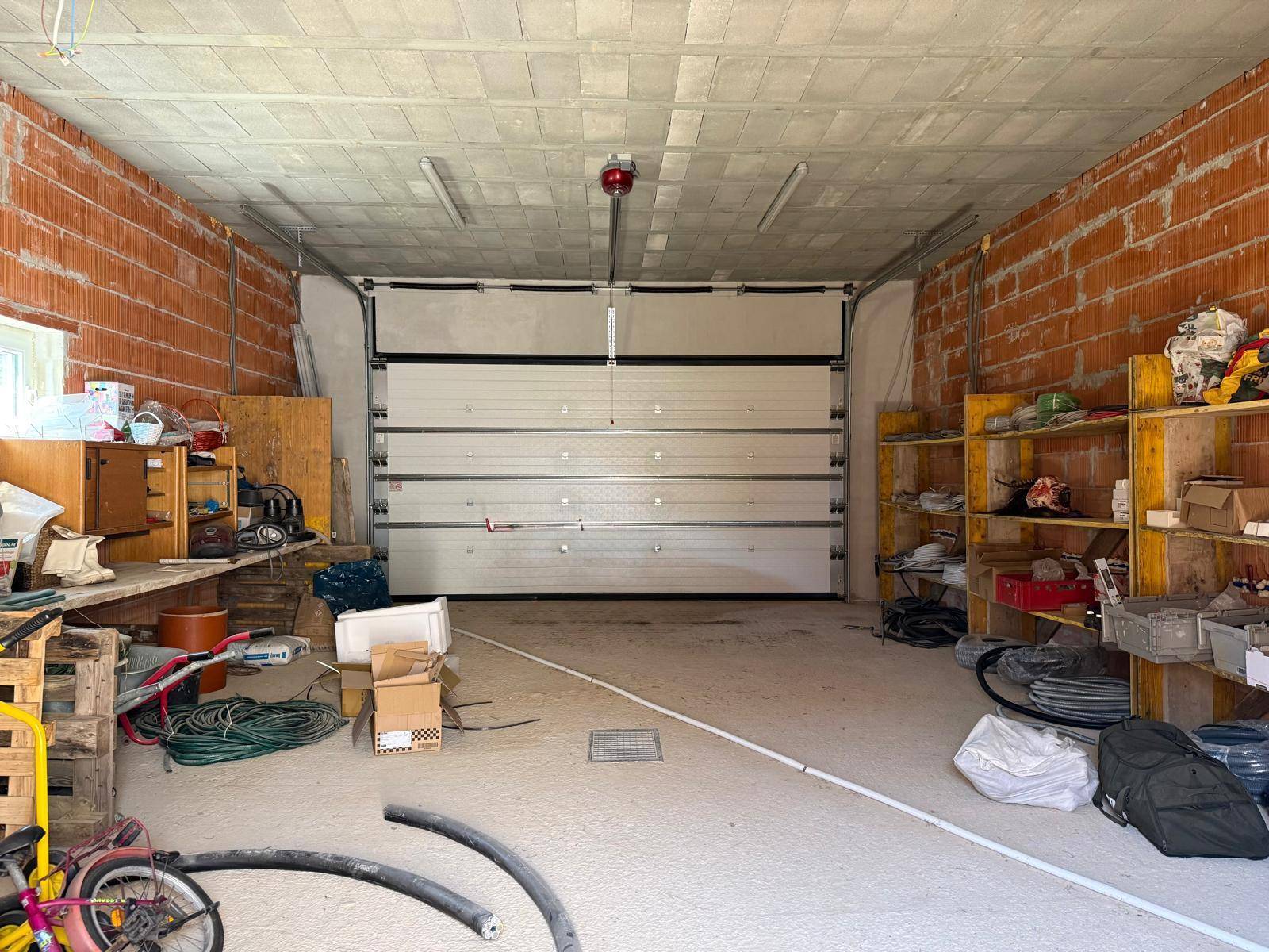 Geräumige Garage im Rohbau mit großem Sektionaltor, Ziegelwänden und provisorischen Regalen für Materialien.