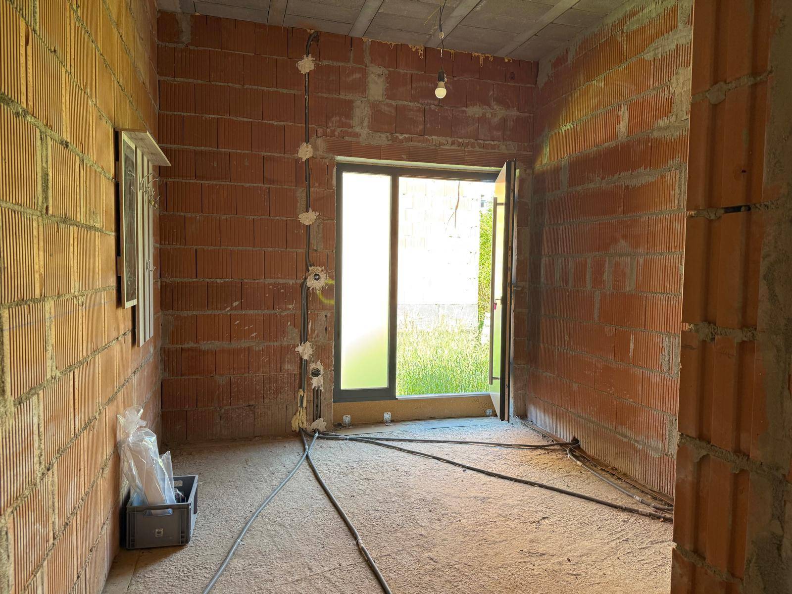 Innenraum im Rohbau mit unverputzten Ziegelwänden, sichtbaren Elektroinstallationen und großem Fenster.