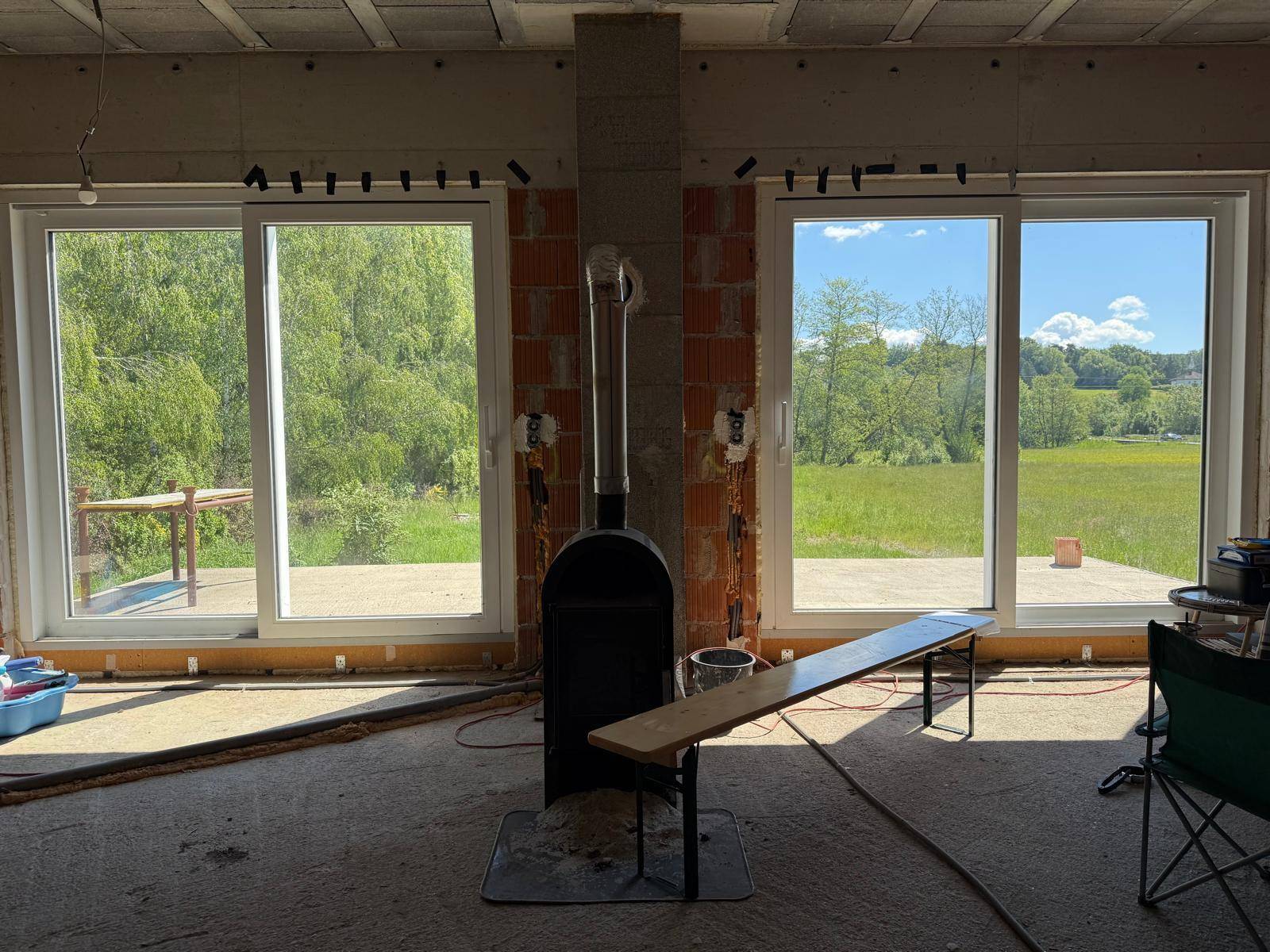 Wohnraum im Rohbau mit großen Fenstern, die einen weiten Blick ins Grüne bieten, und einem Holzofen.