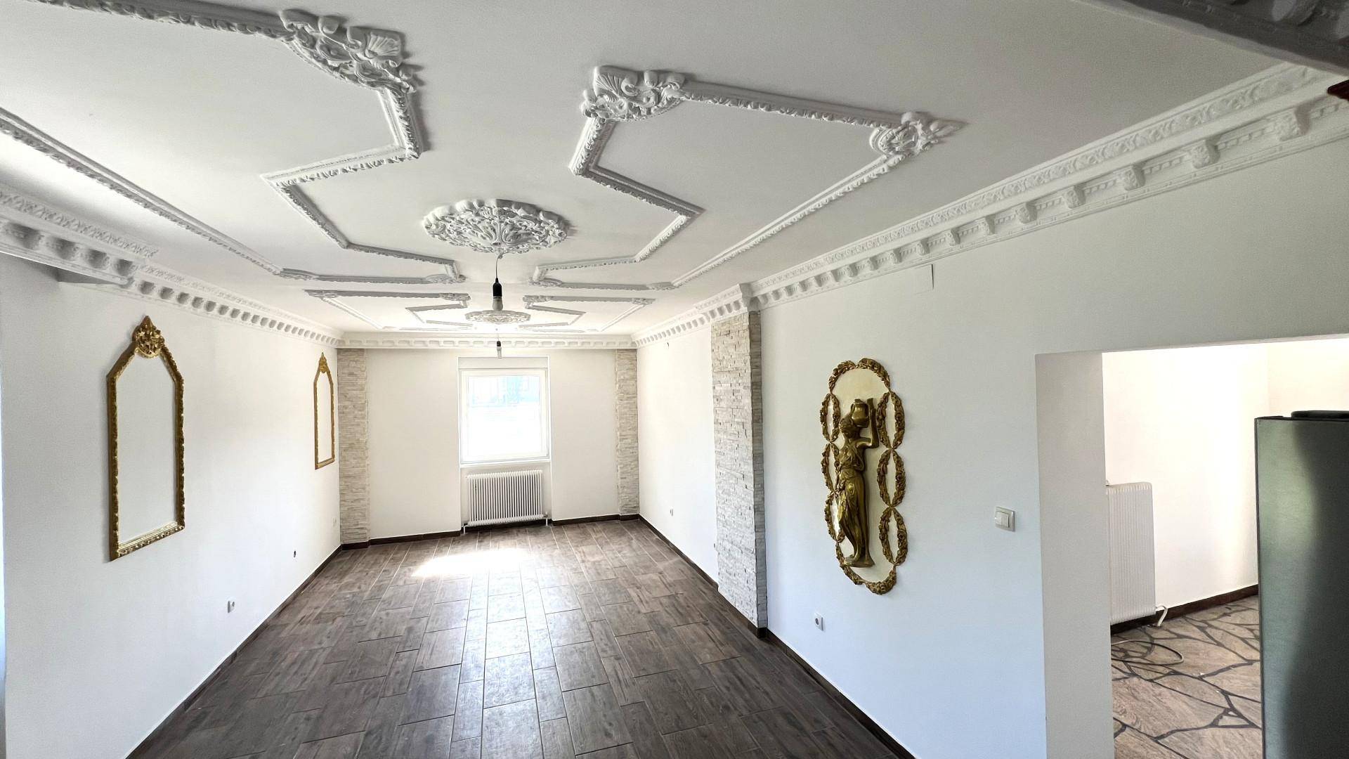 Geräumiges Wohnzimmer mit aufwendiger Stuckdecke, dunklem Boden und einem Fenster.