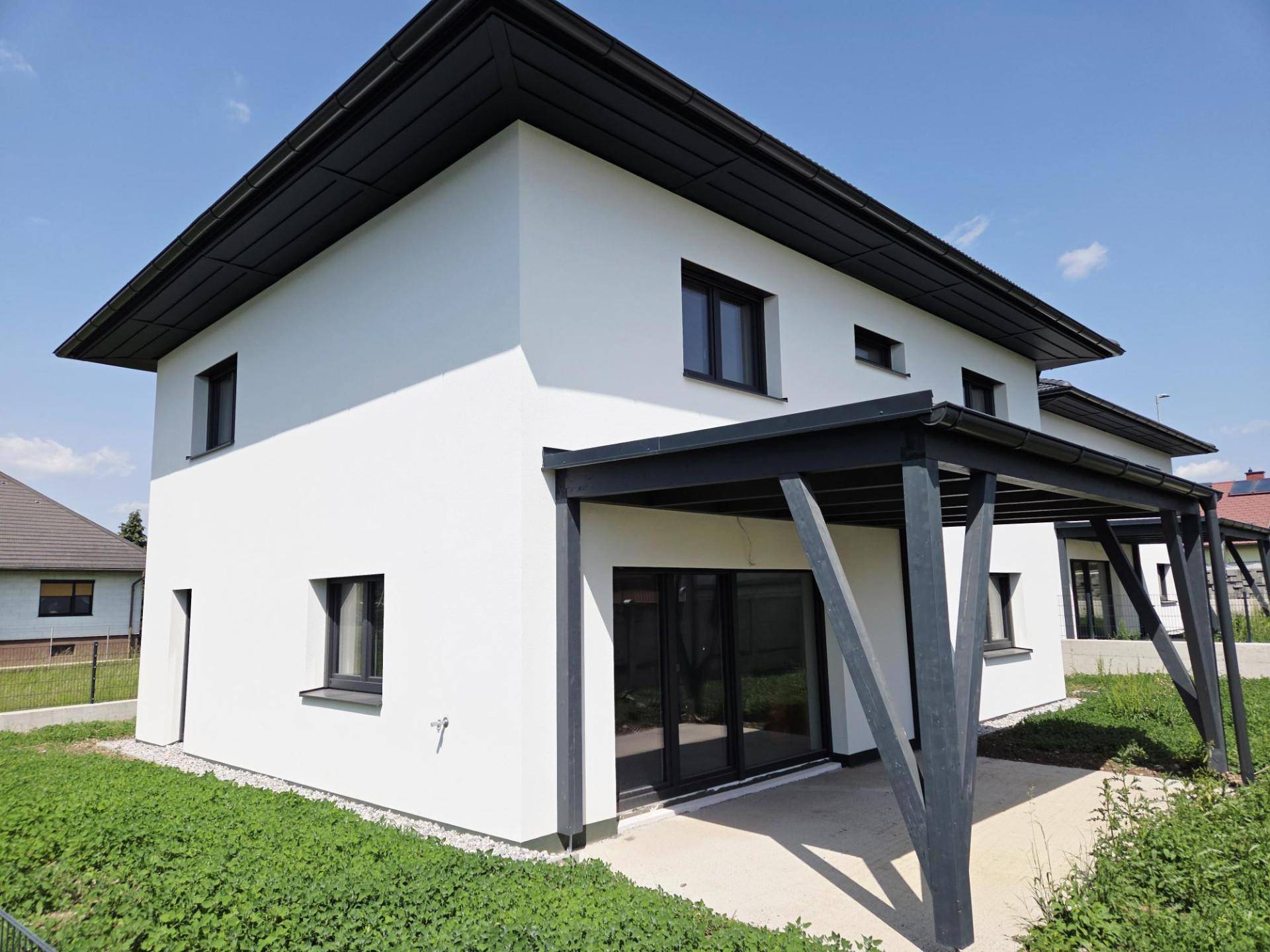 Modernes Haus mit überdachter Terrasse, großen Glasfronten und Gartenblick an einem sonnigen Tag.