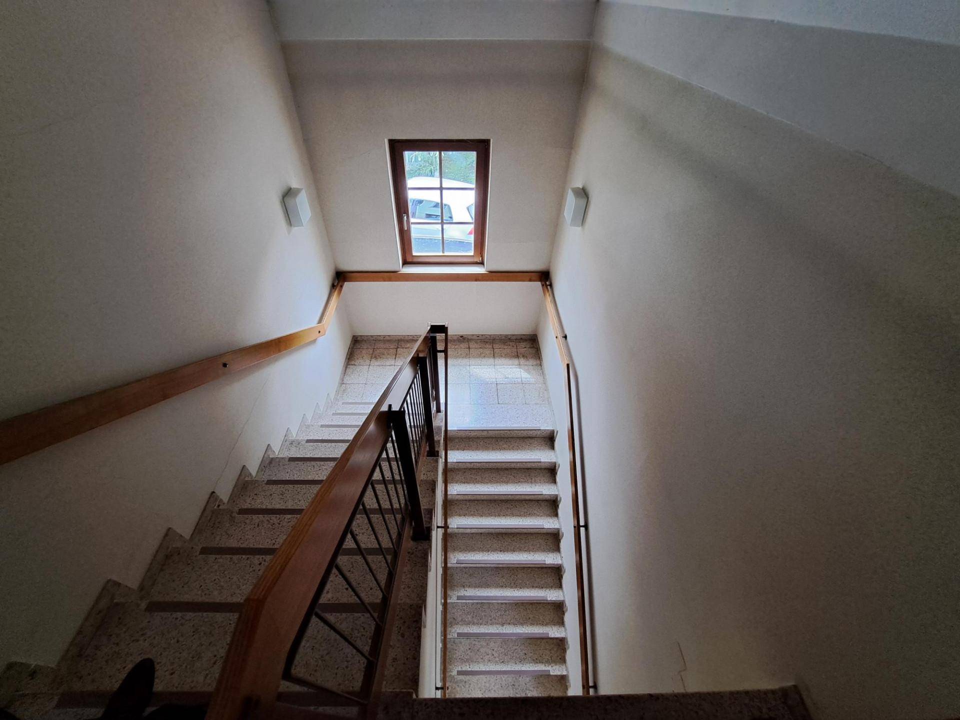 Treppenhaus mit Terrazzoboden, Holzgeländer und Dachfenster für natürliches Licht.