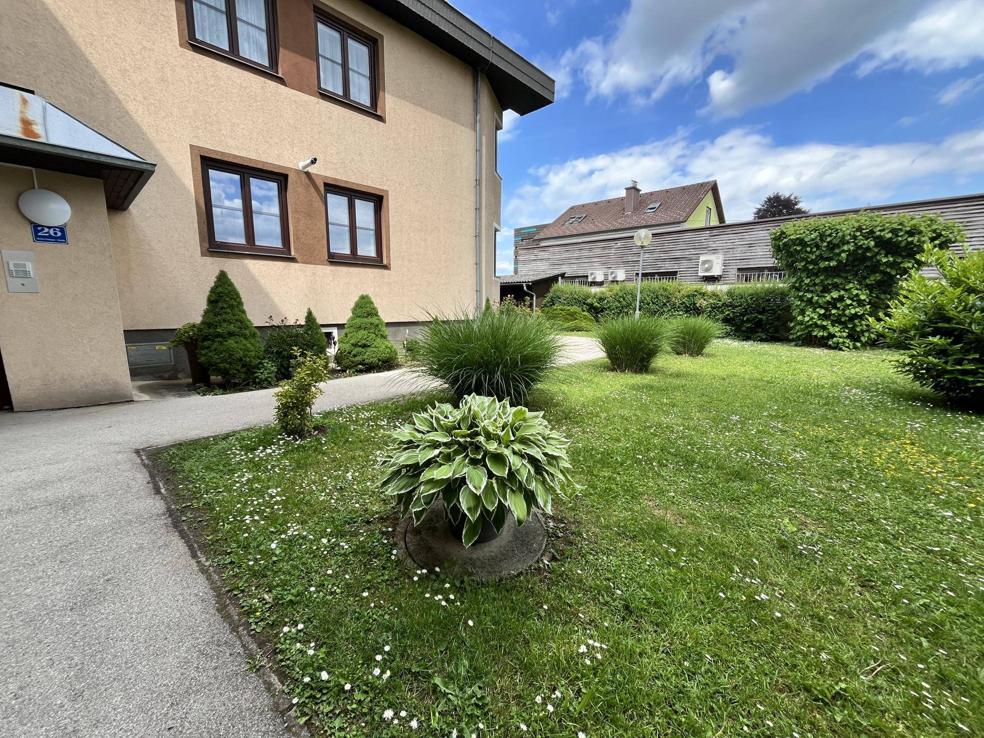 Gepflegte Rasenfläche mit Ziersträuchern vor der Gebäudefassade unter einem bewölkten Himmel.