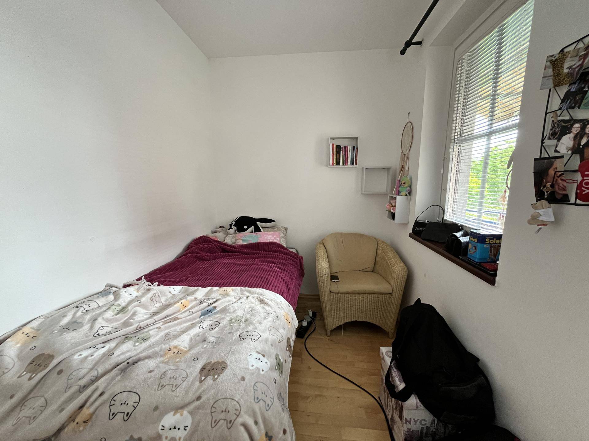 Kleines Schlafzimmer mit Einzelbett, Korbsessel und Fenster mit Jalousien für Privatsphäre.