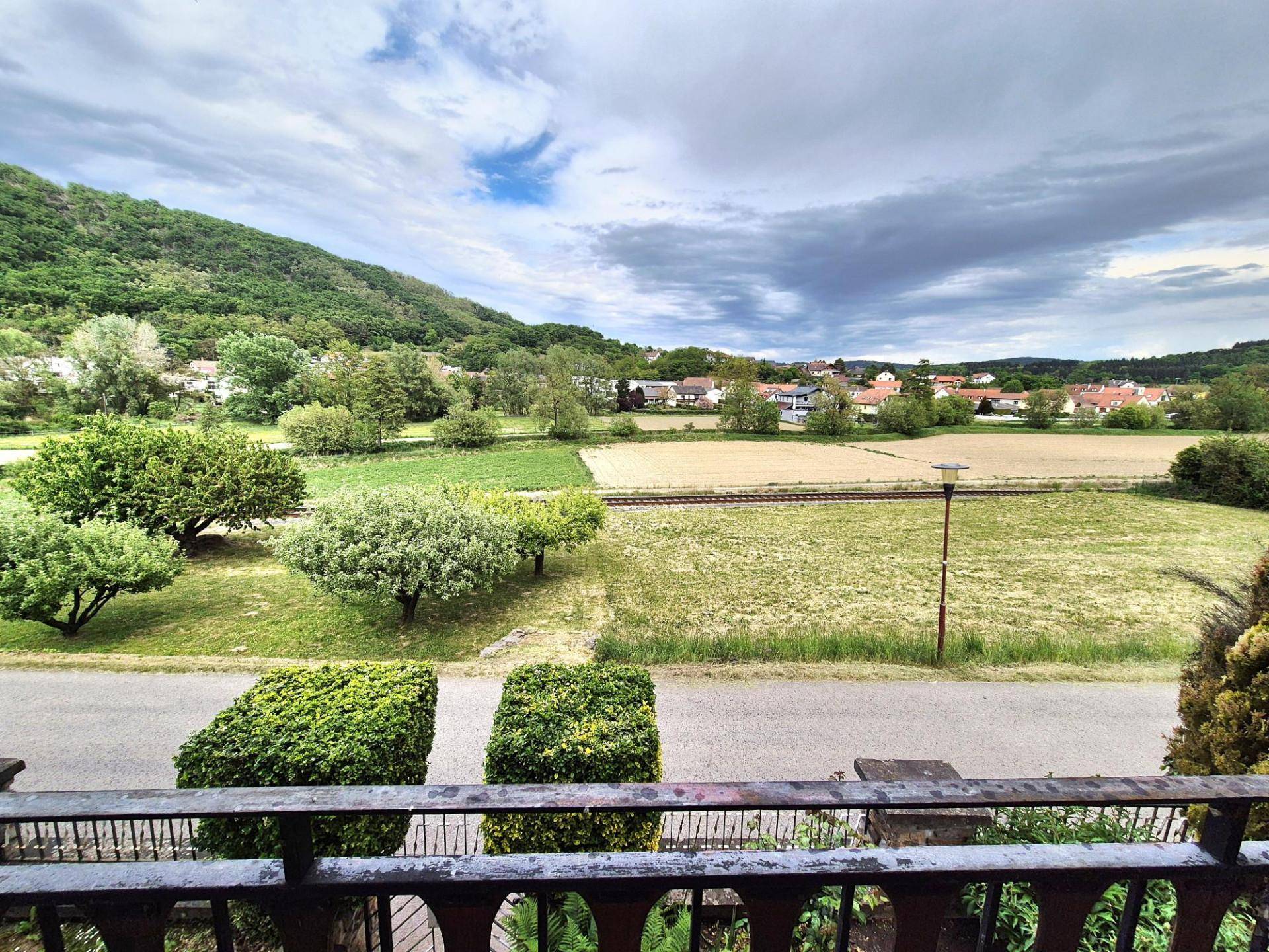 Panoramablick vom Balkon auf grüne Hügel, weite Felder und eine ländliche Siedlung unter bewölktem Himmel.