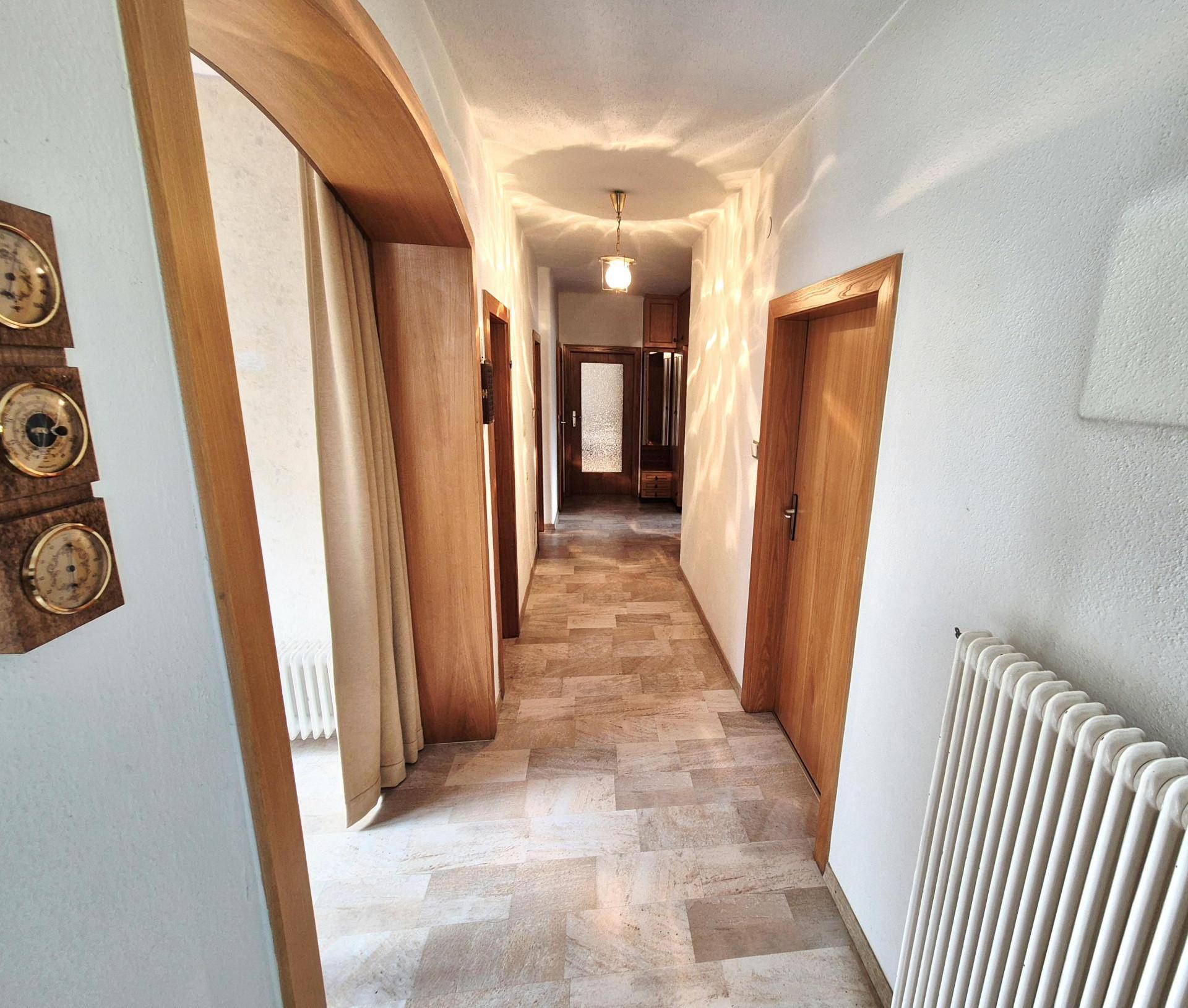 Langer Flur mit hellen Fliesen, Holztüren und einem Heizkörper an der Wand.