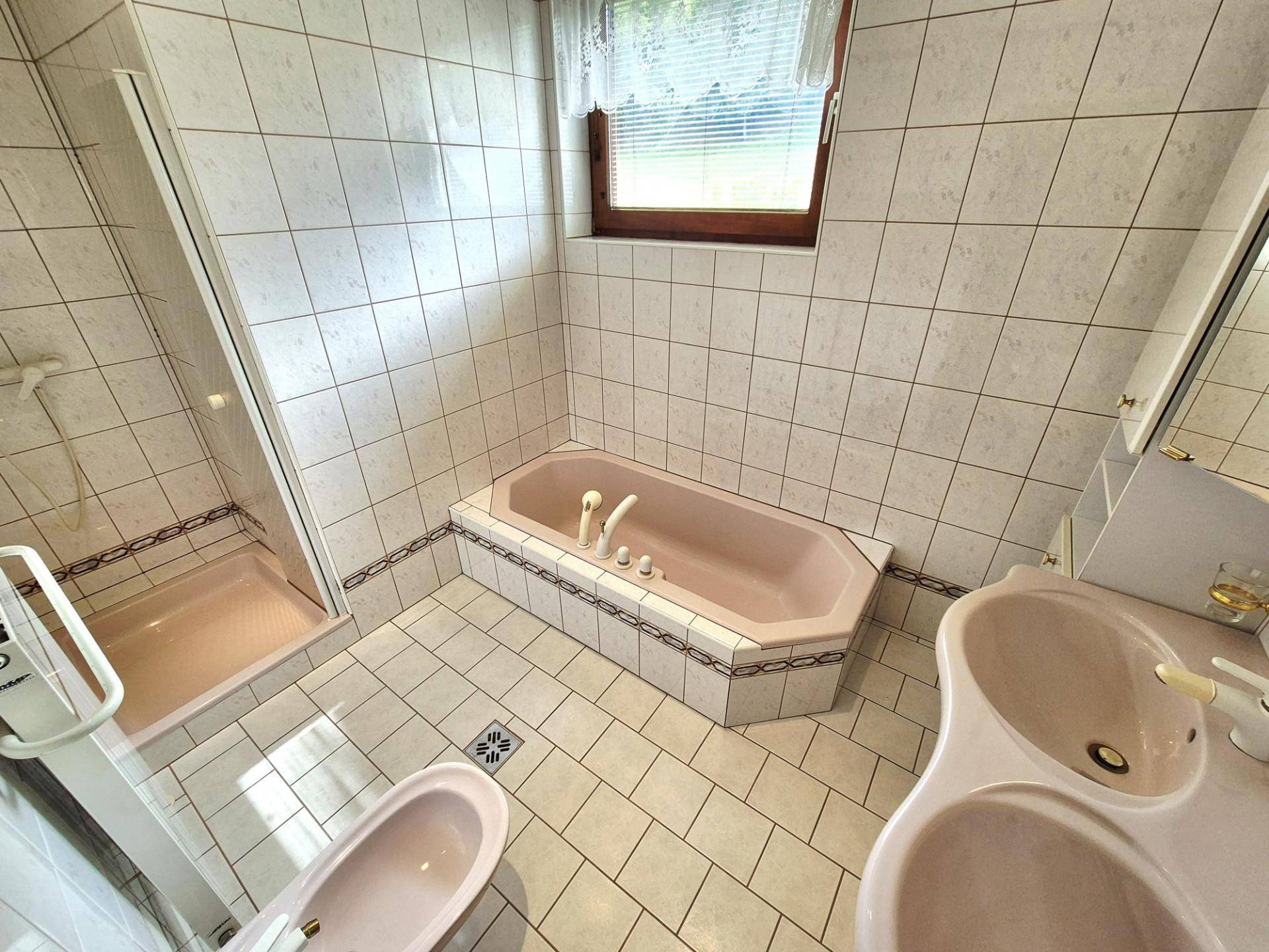 Badezimmer mit rosa Badewanne, Dusche und WC, umgeben von hellen Fliesen und einem Fenster.