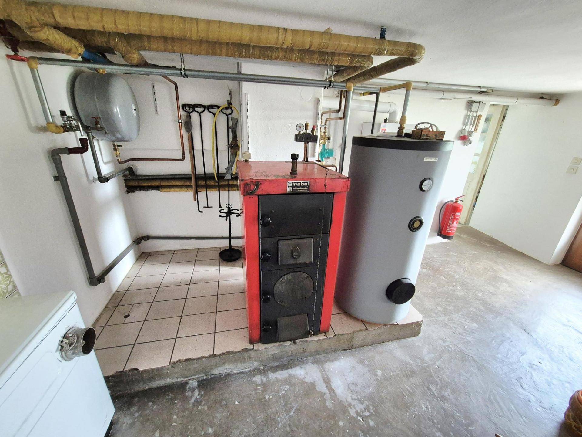 Heizungsraum mit einem älteren Holzofen und Warmwasserspeicher, umgeben von Rohrleitungen und Fliesenboden.