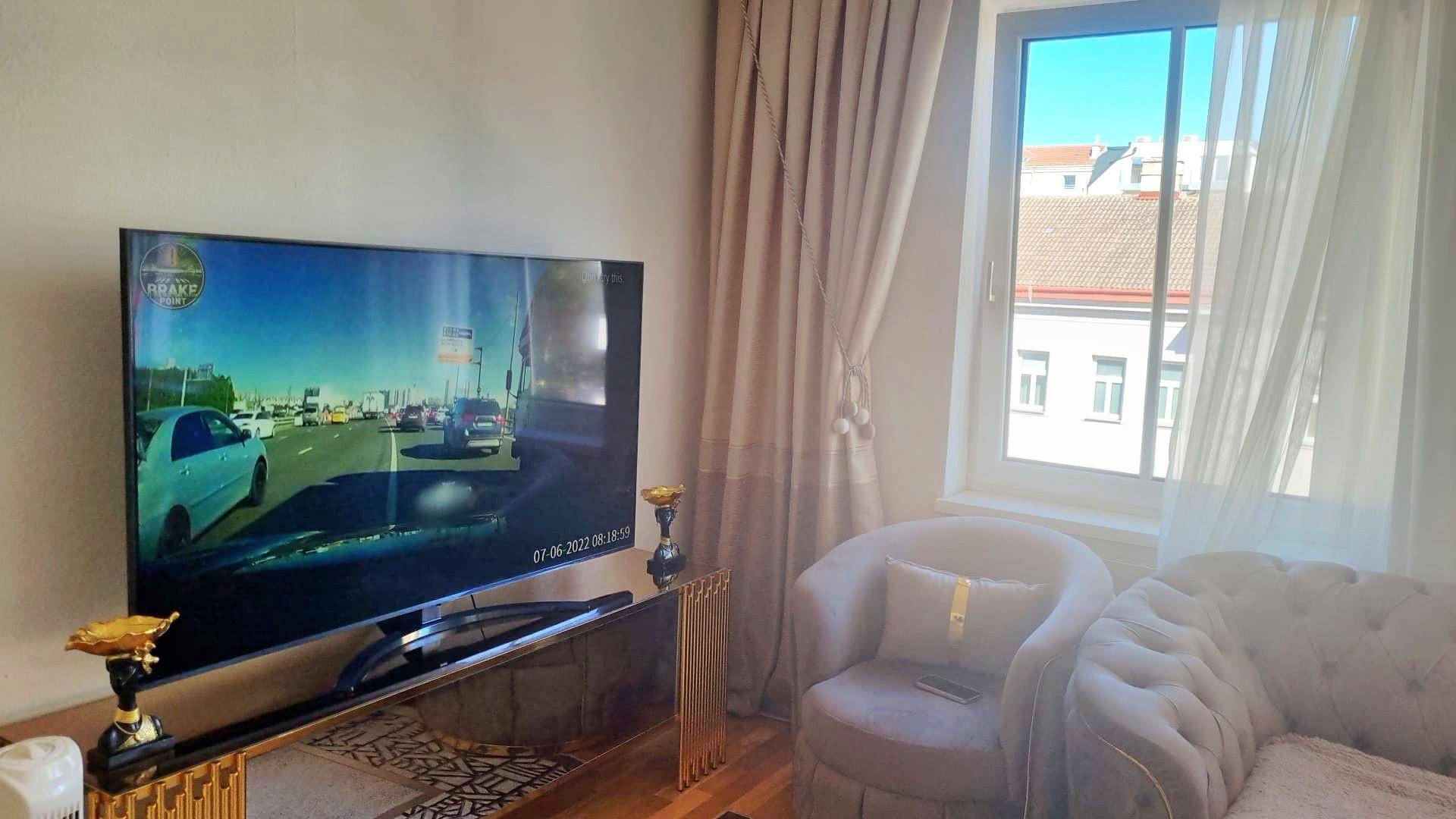 Gemütliches Wohnzimmer mit großem Fernseher, beigen Vorhängen und Blick auf die Stadt.