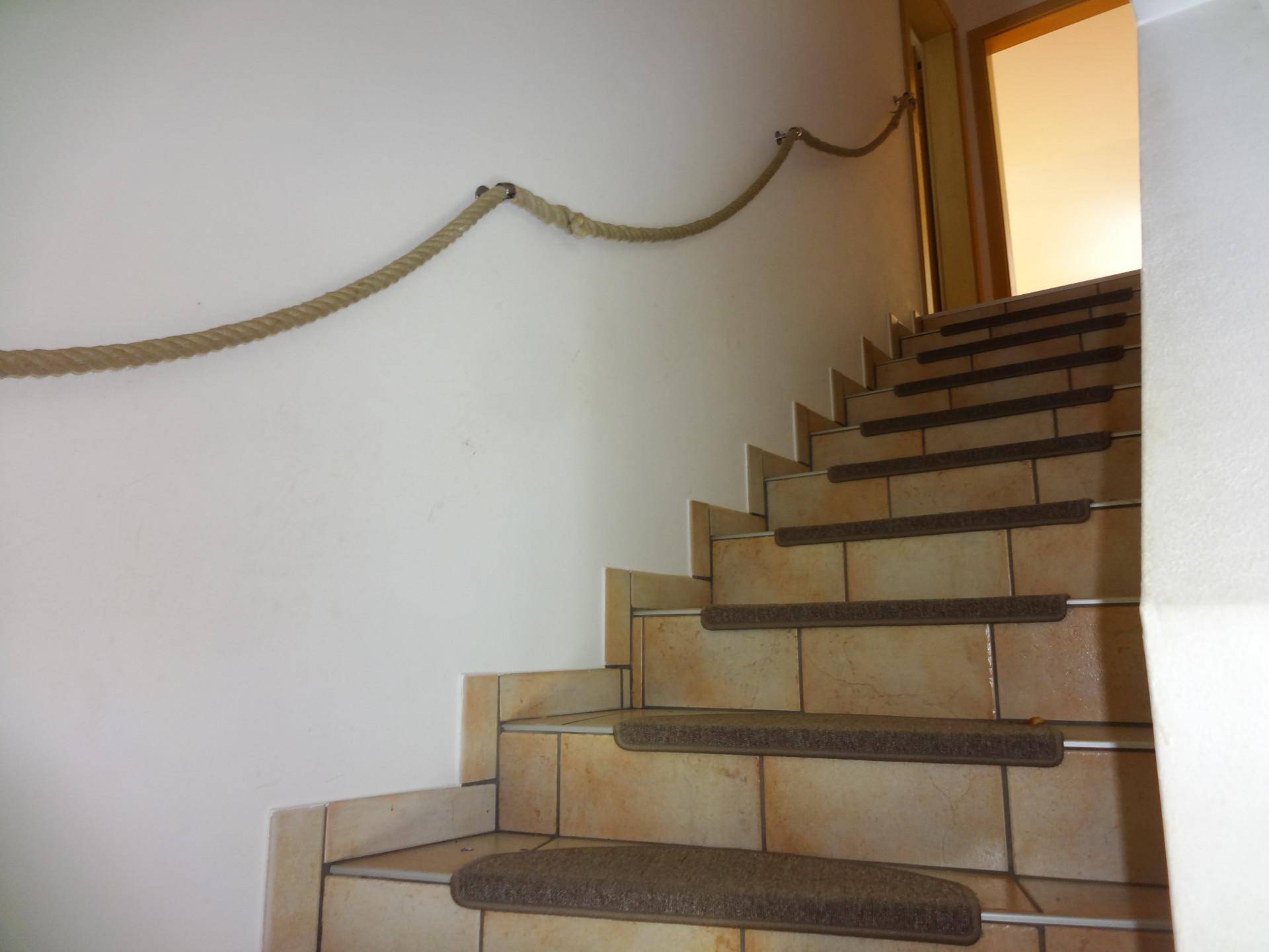 Treppenhaus mit gefliesten Stufen, Teppichläufern und einem Seil als rustikalem Handlauf.