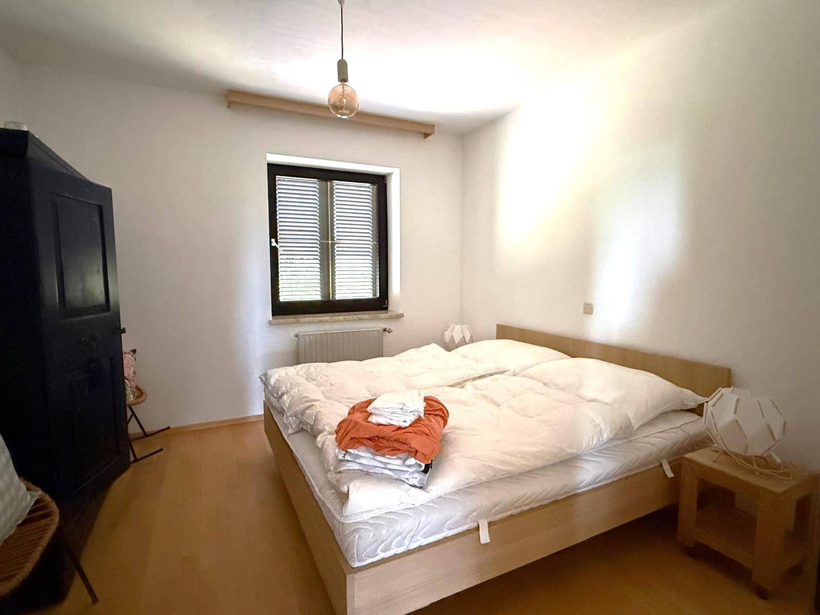 Helles Schlafzimmer mit Holzbett, dunklem Kleiderschrank und Fenster mit Jalousien.