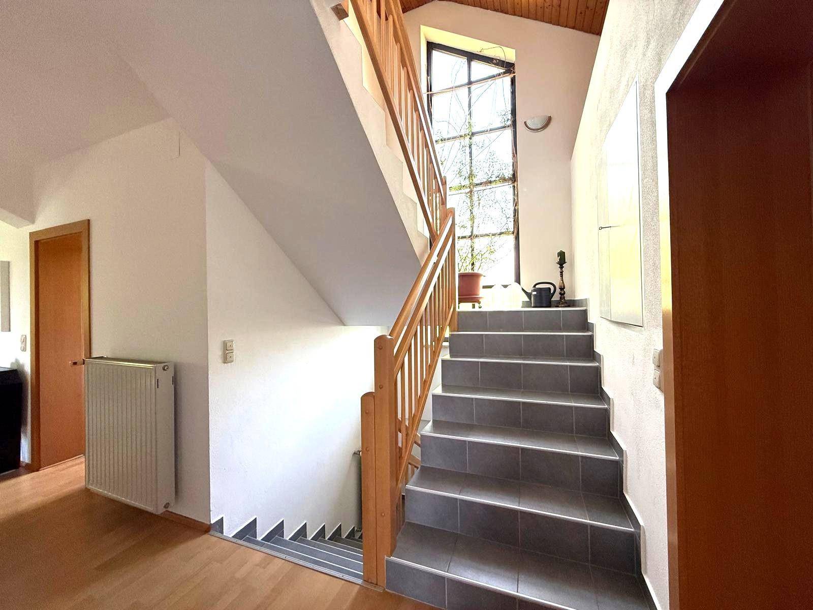 Treppenhaus mit Holzgeländer und einem großen, hohen Fenster, das viel Licht spendet.