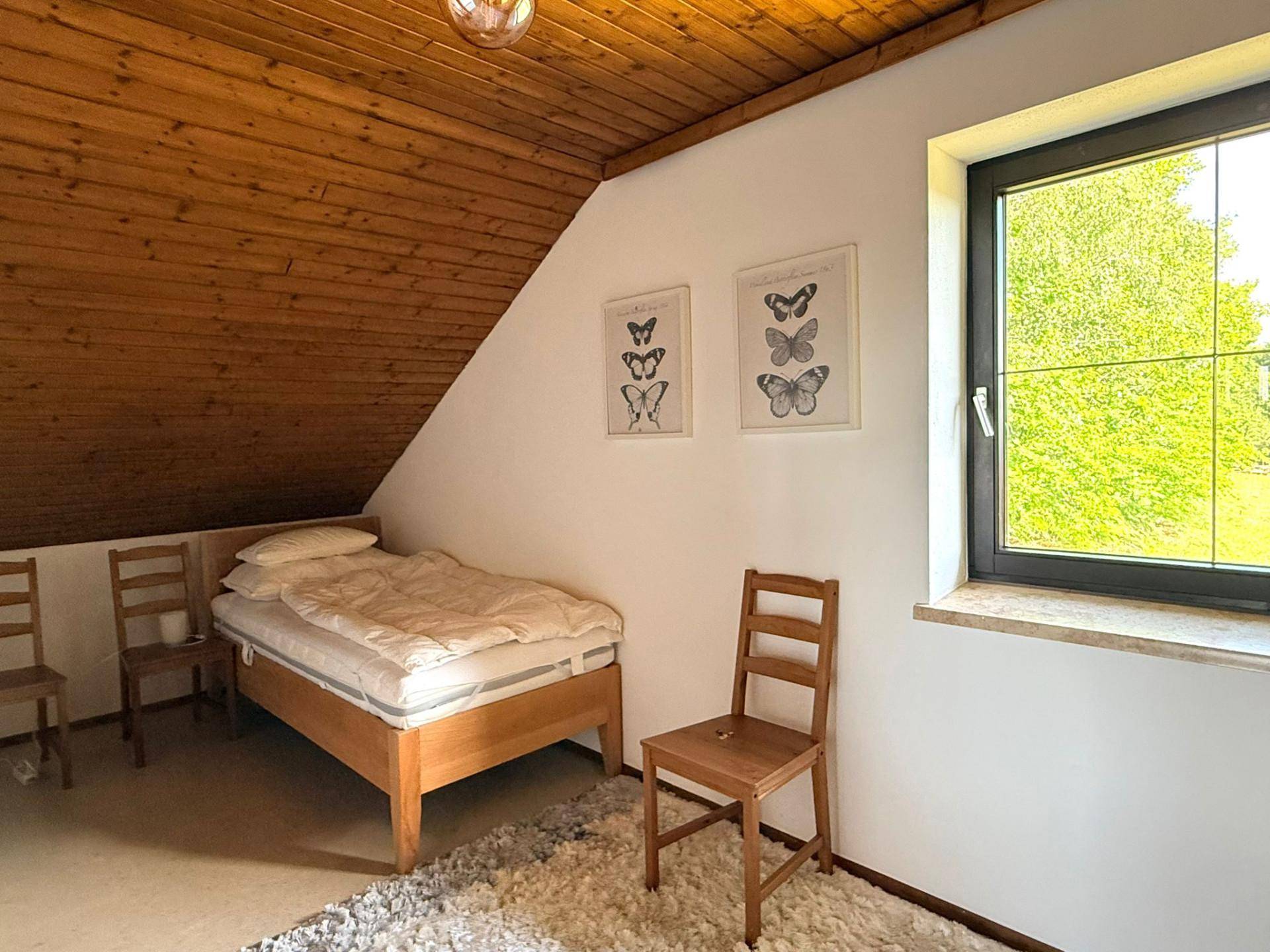 Schlafzimmer im Dachgeschoss mit Holztäfelung, Einzelbett und Fenster mit Ausblick ins Grüne.
