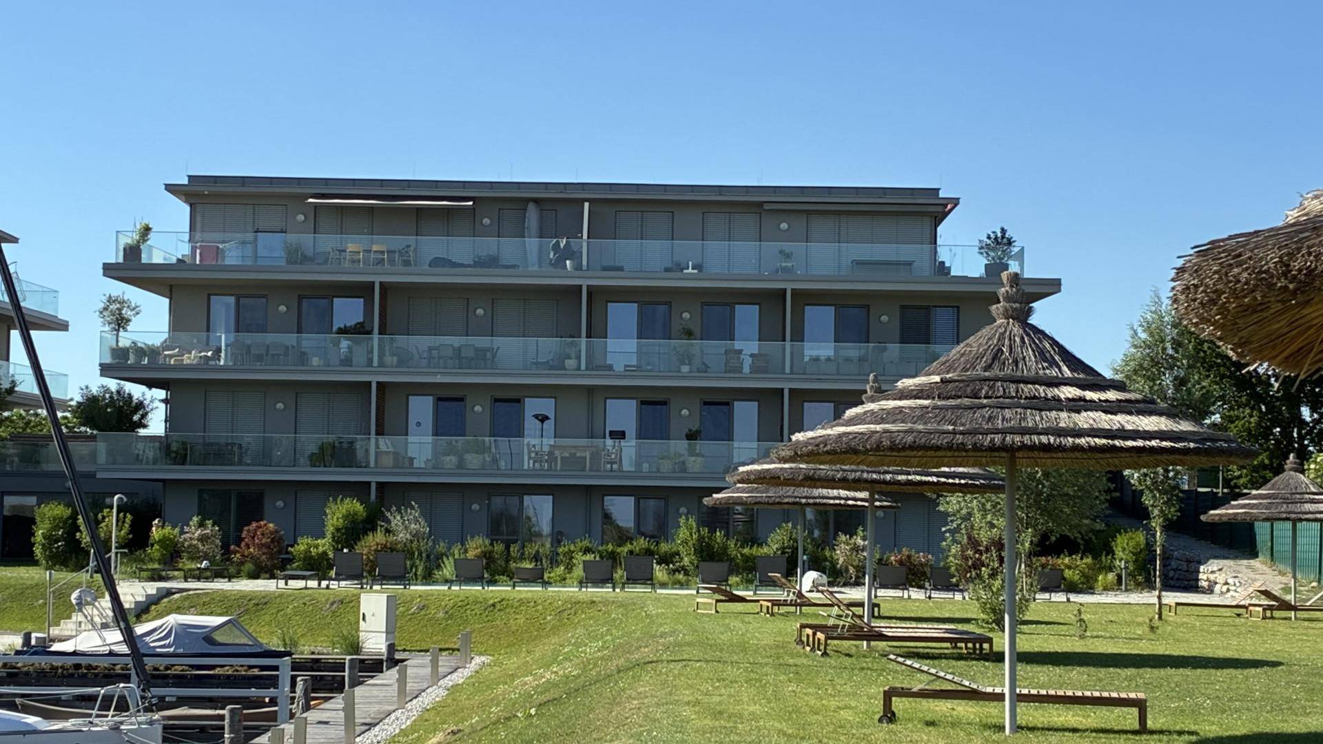 Modernes Apartmentgebäude mit Balkonen, umgeben von einer Liegewiese mit Sonnenschirmen am Seeufer.