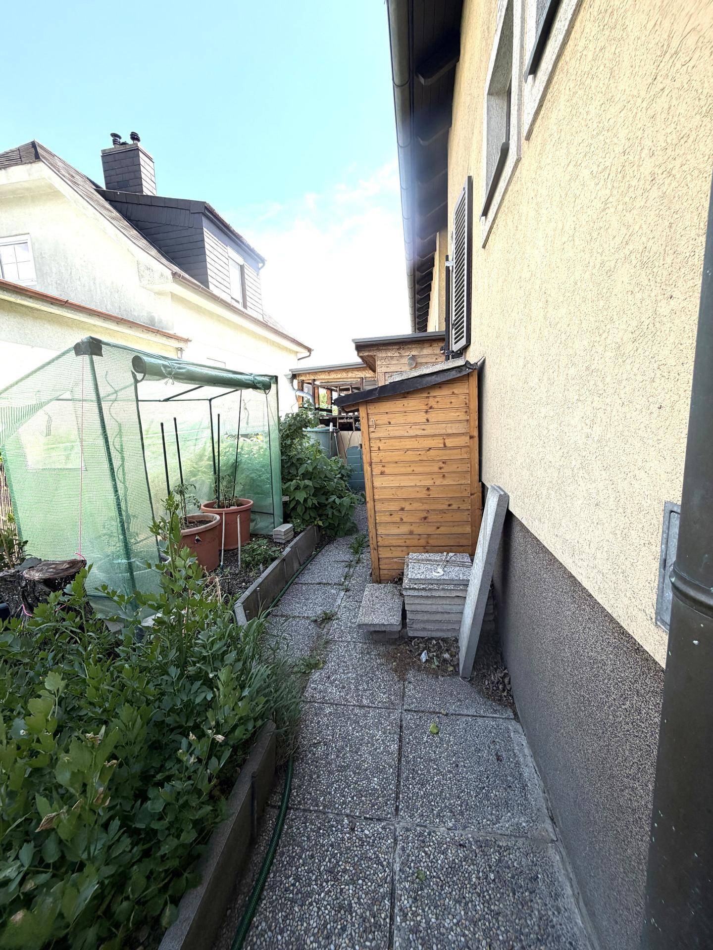 Schmaler Gartenweg neben dem Haus mit Gewächshaus und Geräteschuppen.