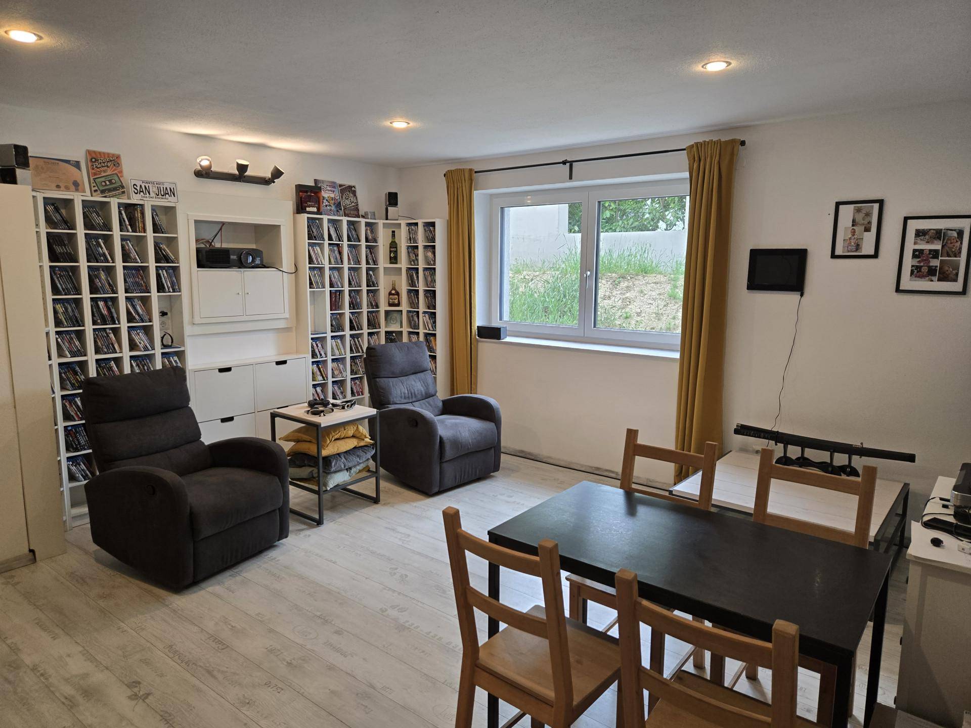 Gemütliches Wohnzimmer mit zwei Relaxsesseln, Esstisch und Regalen voller Medien, ideal für Unterhaltung.