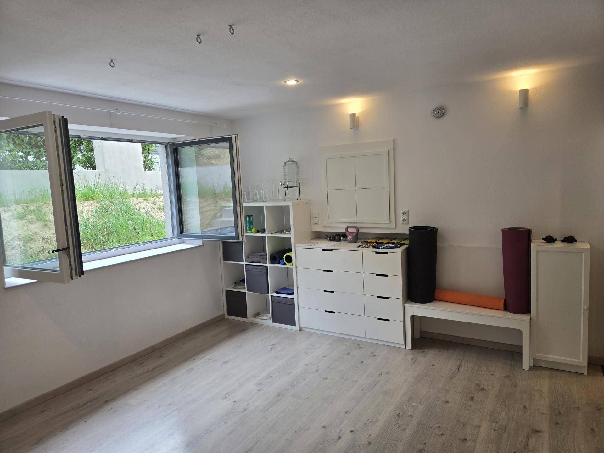 Heller Mehrzweckraum mit großem Fenster, Laminatboden und weißen Möbeln, vielseitig nutzbar.