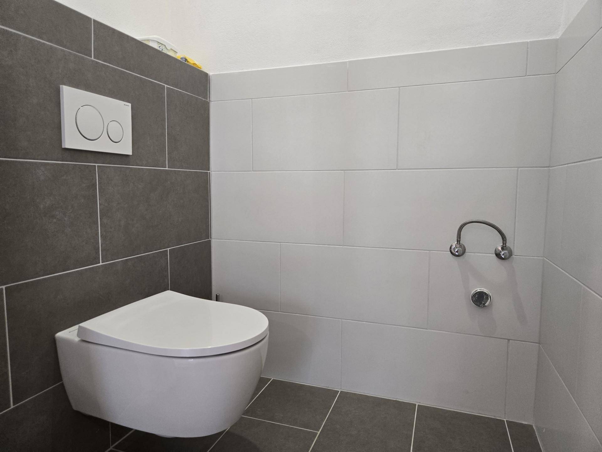 Modernes WC mit wandhängender Toilette und grauen sowie weißen Fliesen an den Wänden.