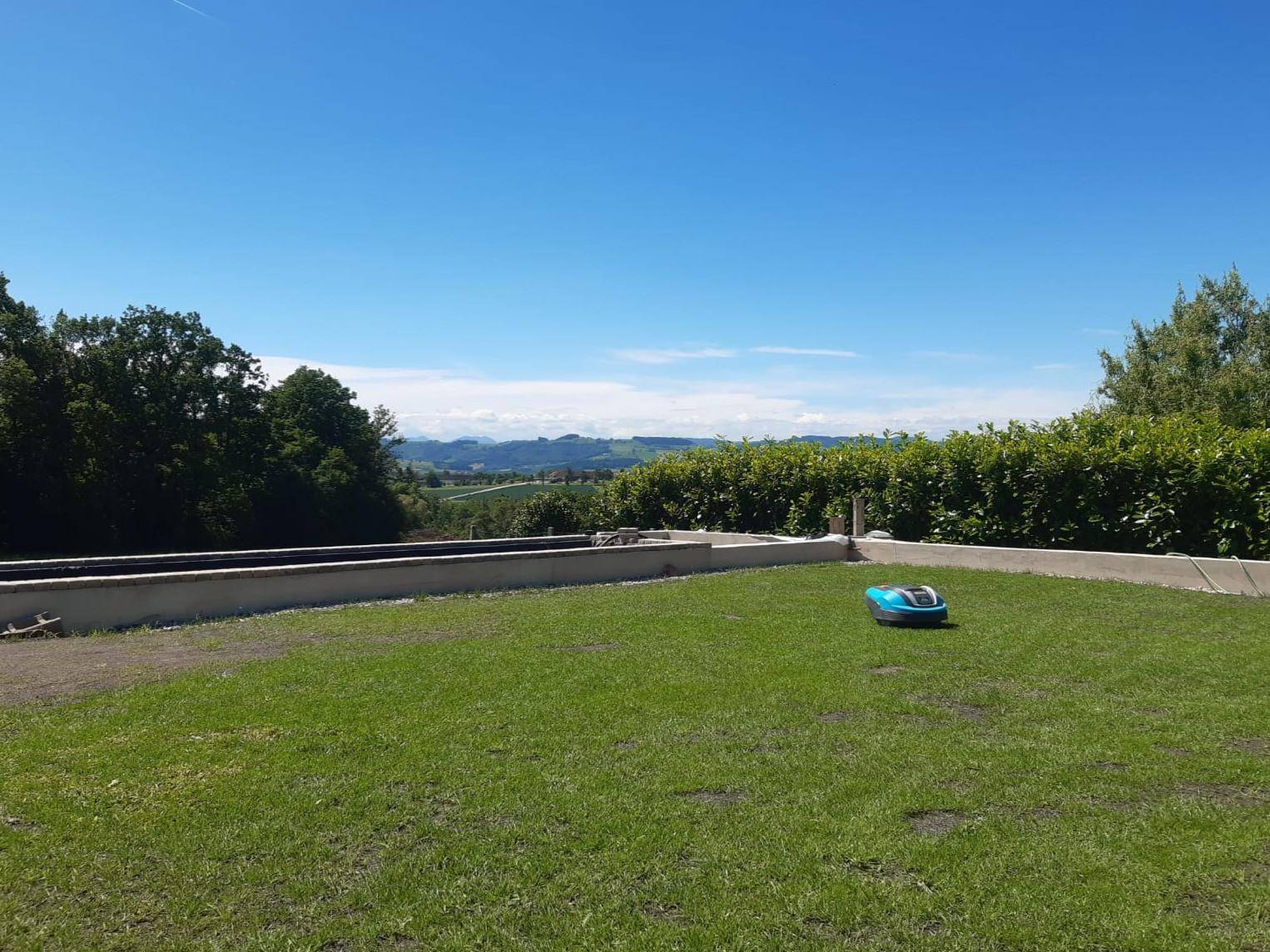 Gepflegter Rasen mit Mähroboter und weitem Blick über die grüne Landschaft unter blauem Himmel.